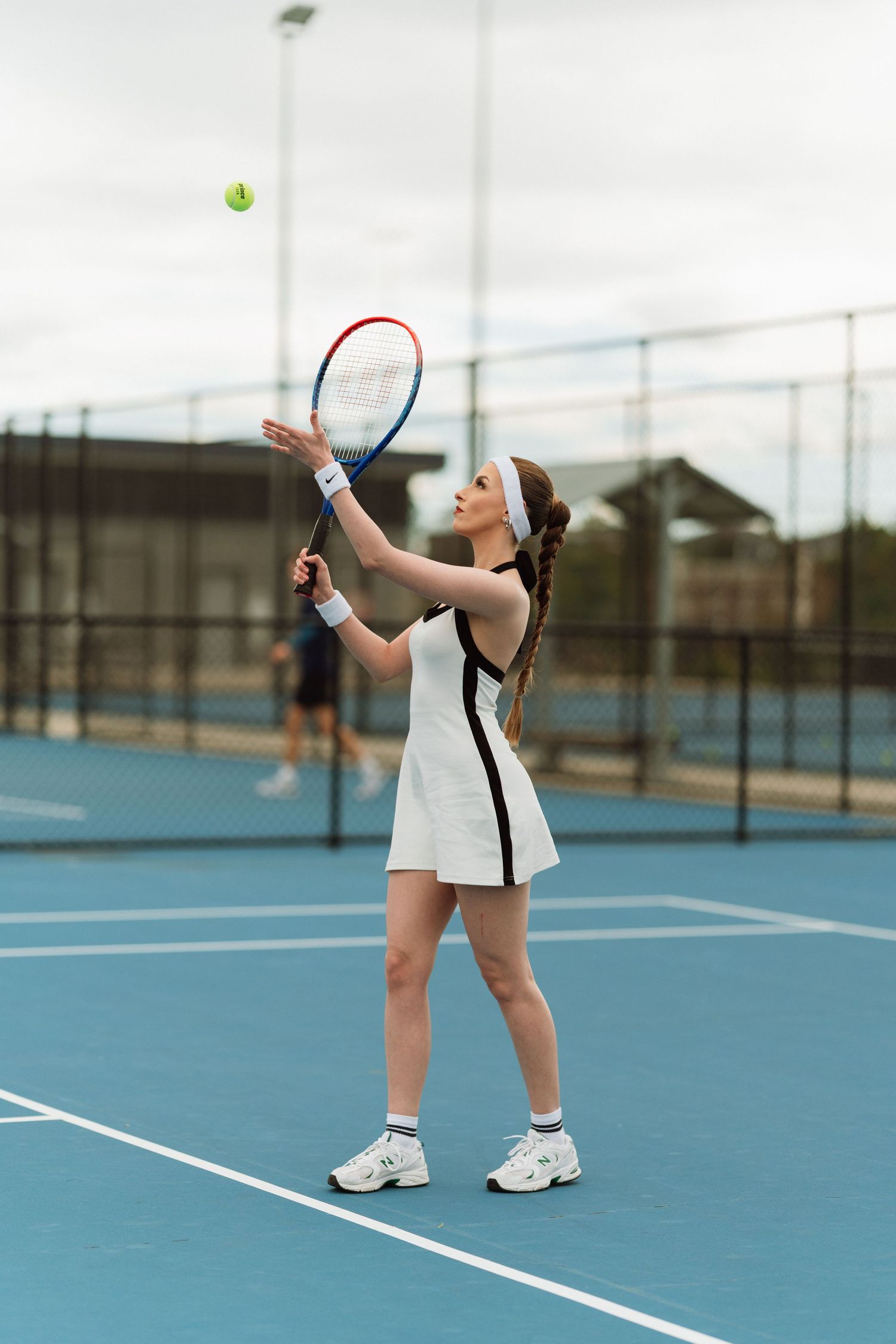 Woman in white tennis dress tossing ball to serve on blue outdoor tennis court.