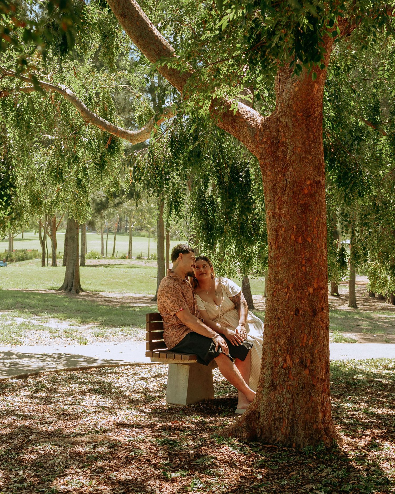 Couple sitting together on a bench under a large tree in a sunny green park.