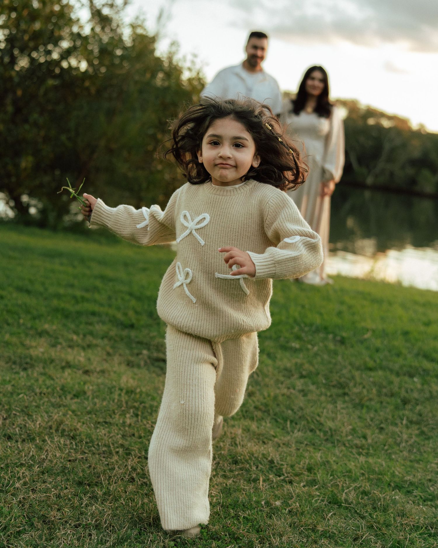 A joyful curly-haired girl in a beige knit outfit runs toward the camera while her parents smile in the background near a lake.