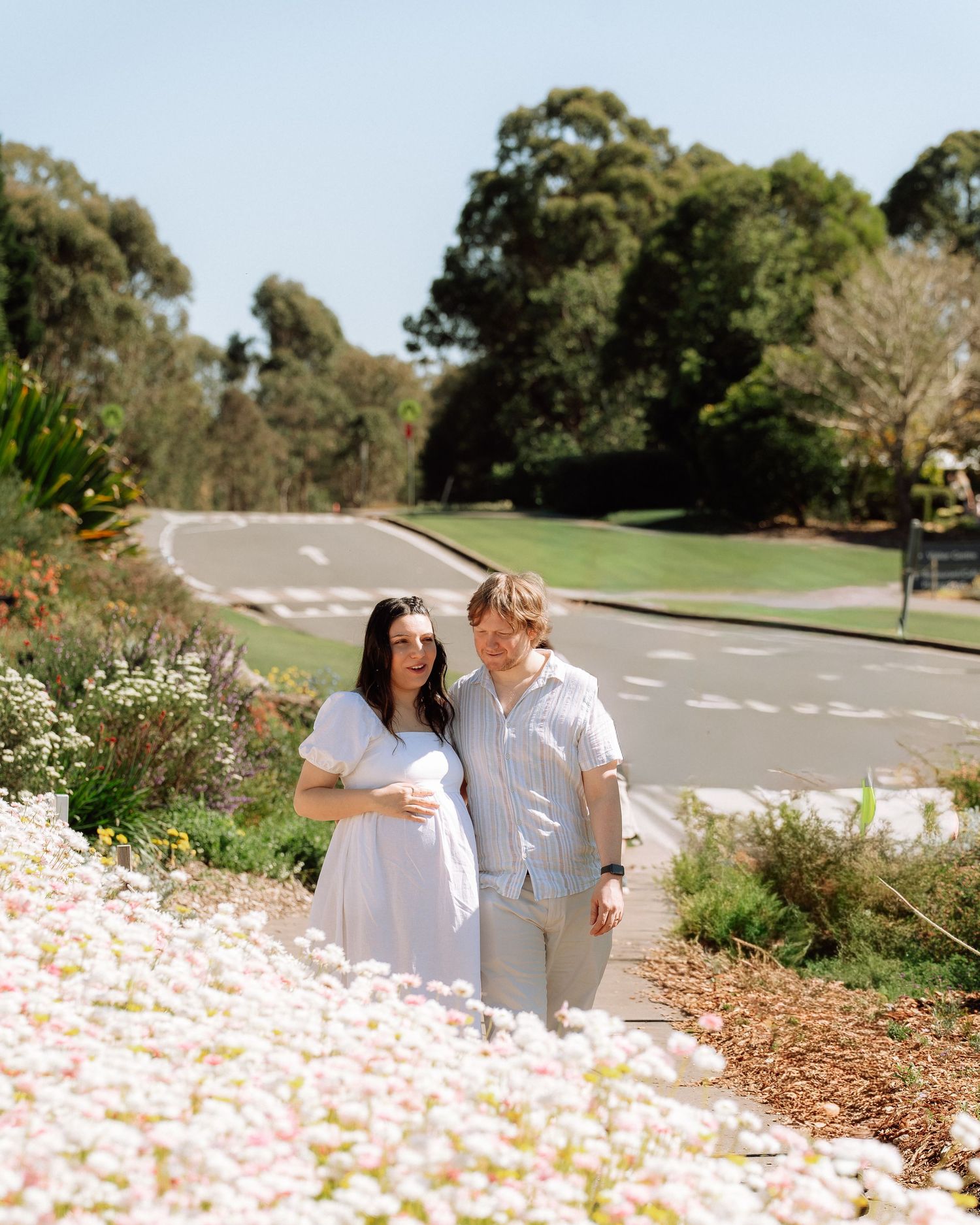 Pregnant couple standing among blooming white flowers in a sunny botanical garden setting.