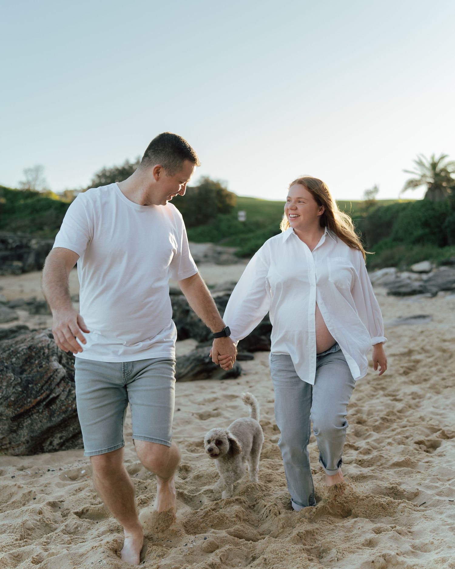A couple holding hands and walking barefoot on a sandy beach with a small dog, wearing white shirts and gray pants.