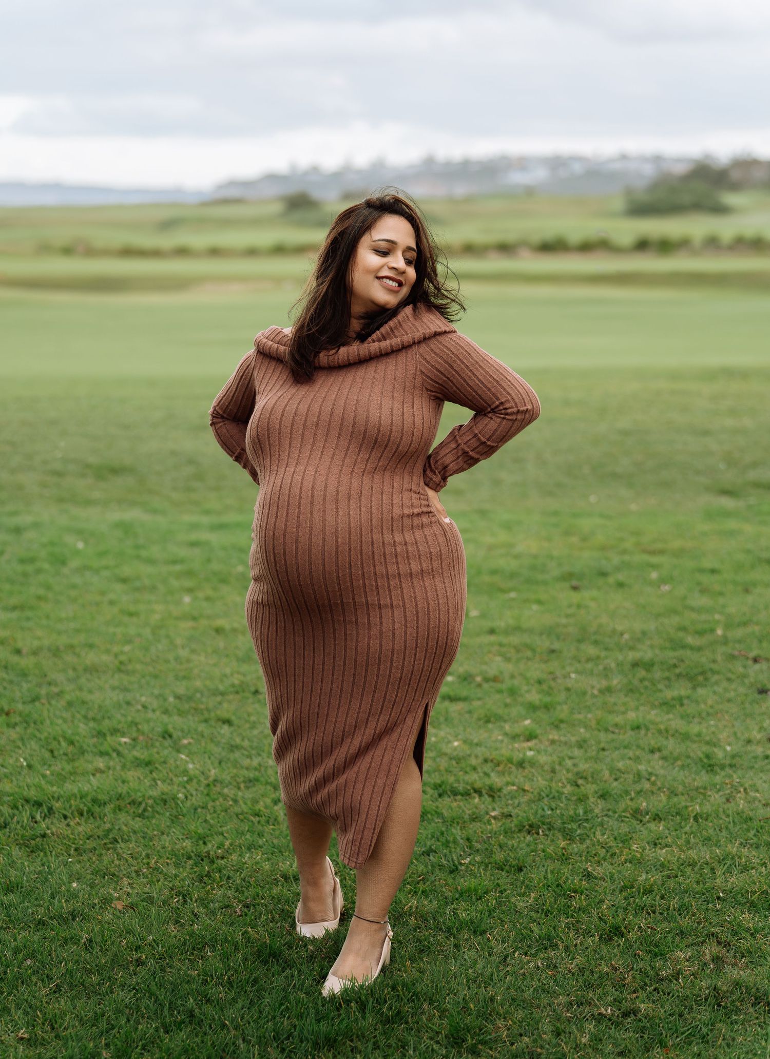 Pregnant woman in brown ribbed turtleneck midi dress posing on green grass field with scenic countryside backdrop.