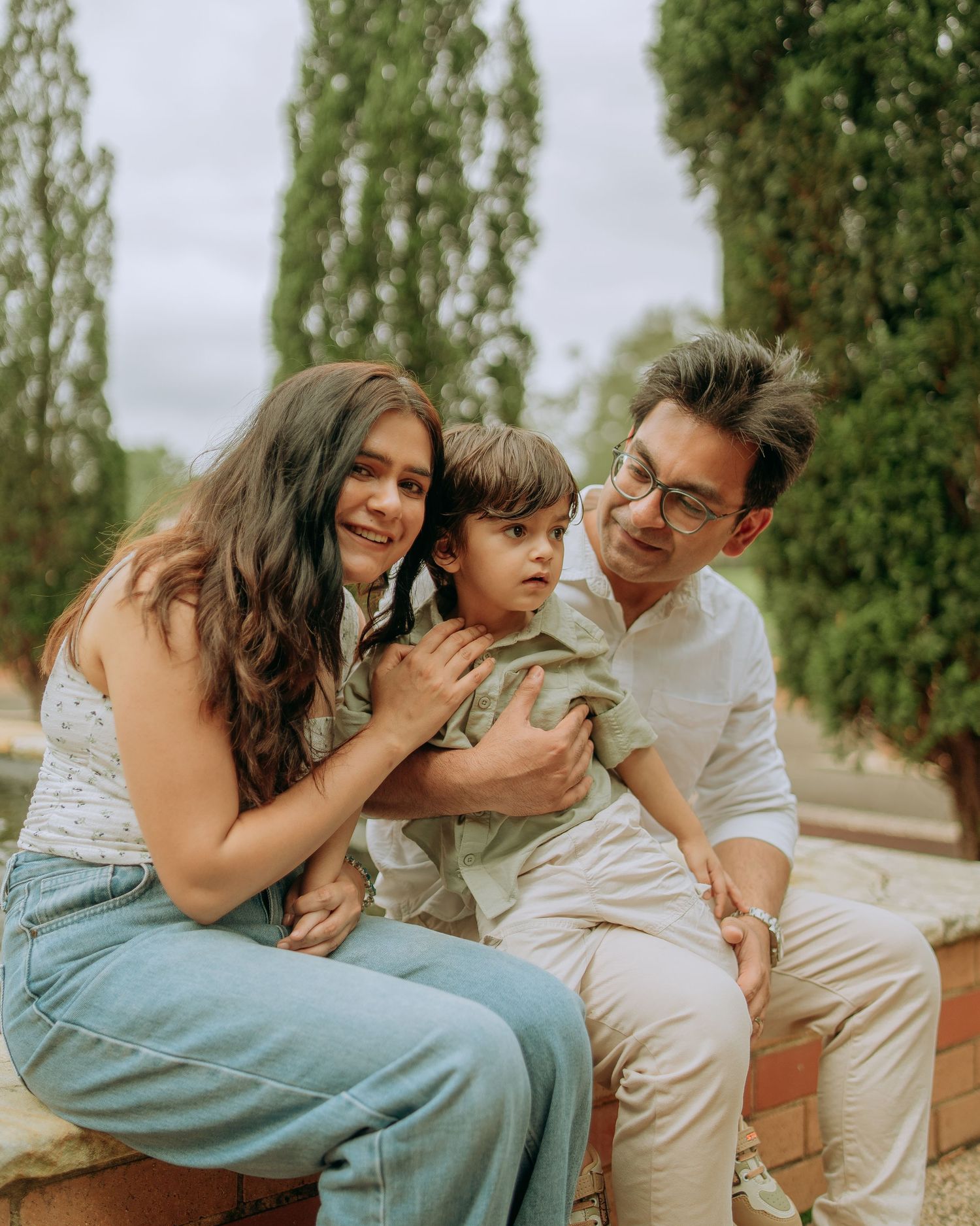 Family of three sharing a joyful moment on brick wall, parents embracing young child in tree-lined garden.