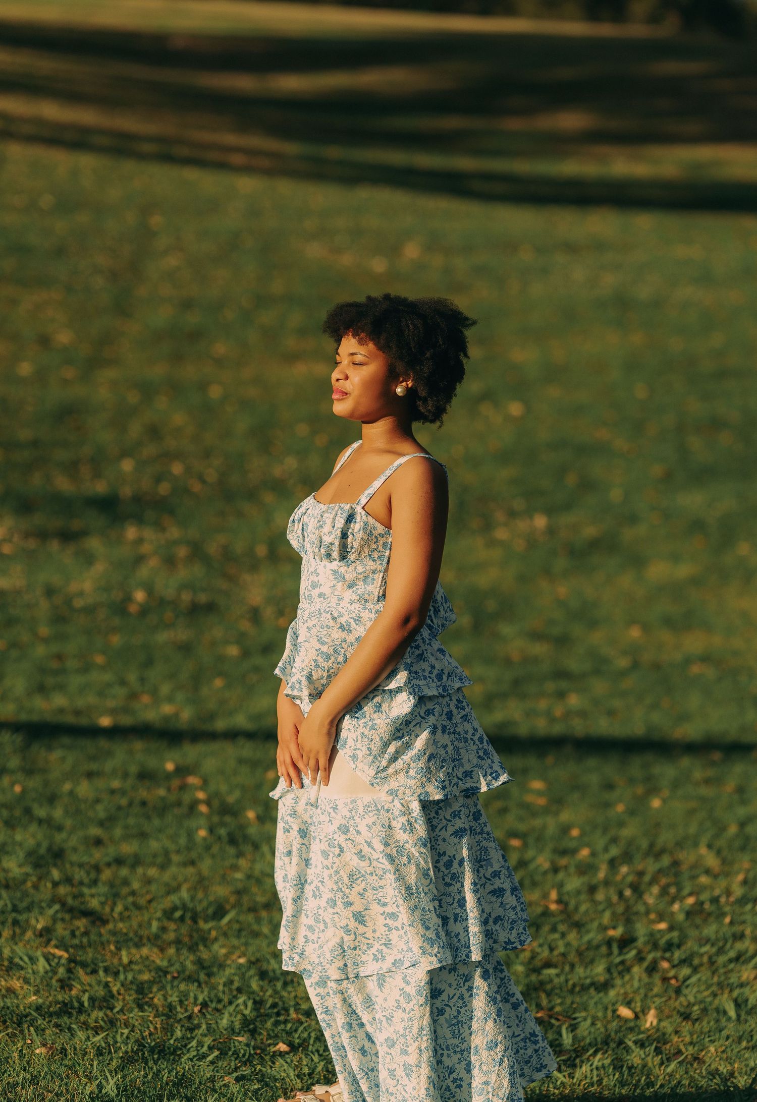 A young woman in a blue floral tiered dress stands peacefully in a sunlit green park with eyes closed.
