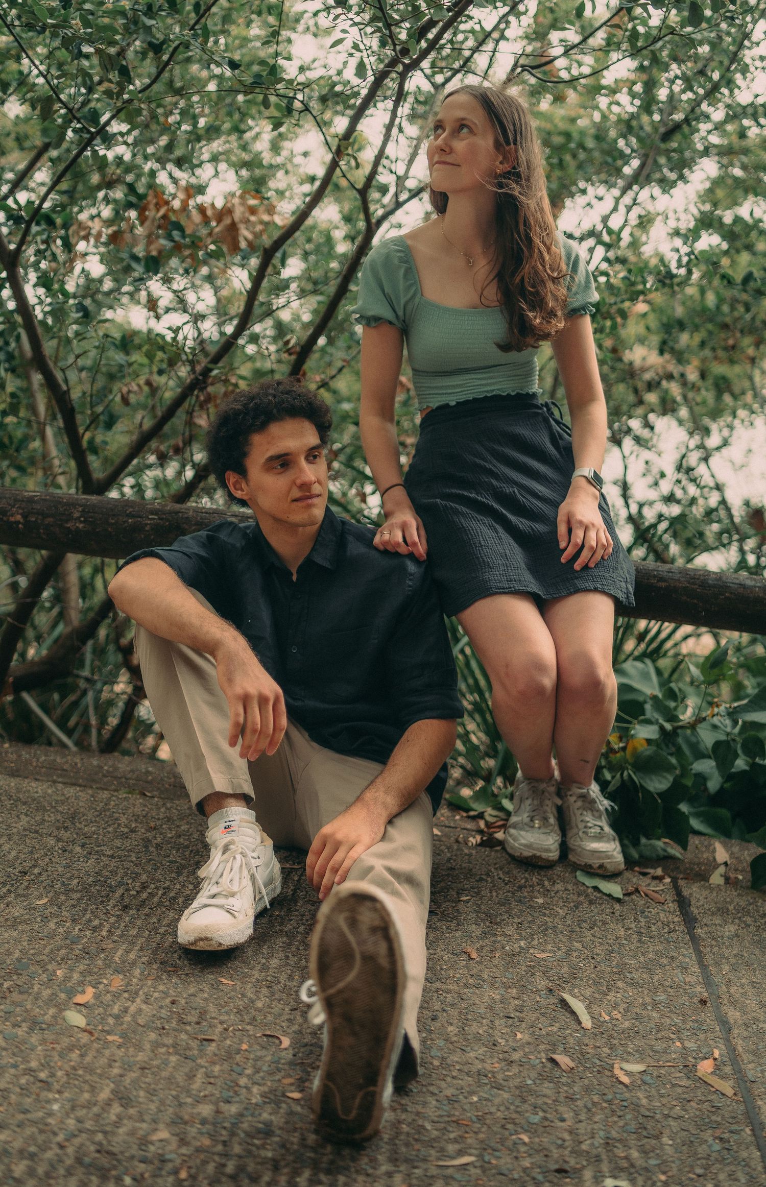 Young couple posing outdoors, man sitting on ground while woman perches on a wooden railing among trees.