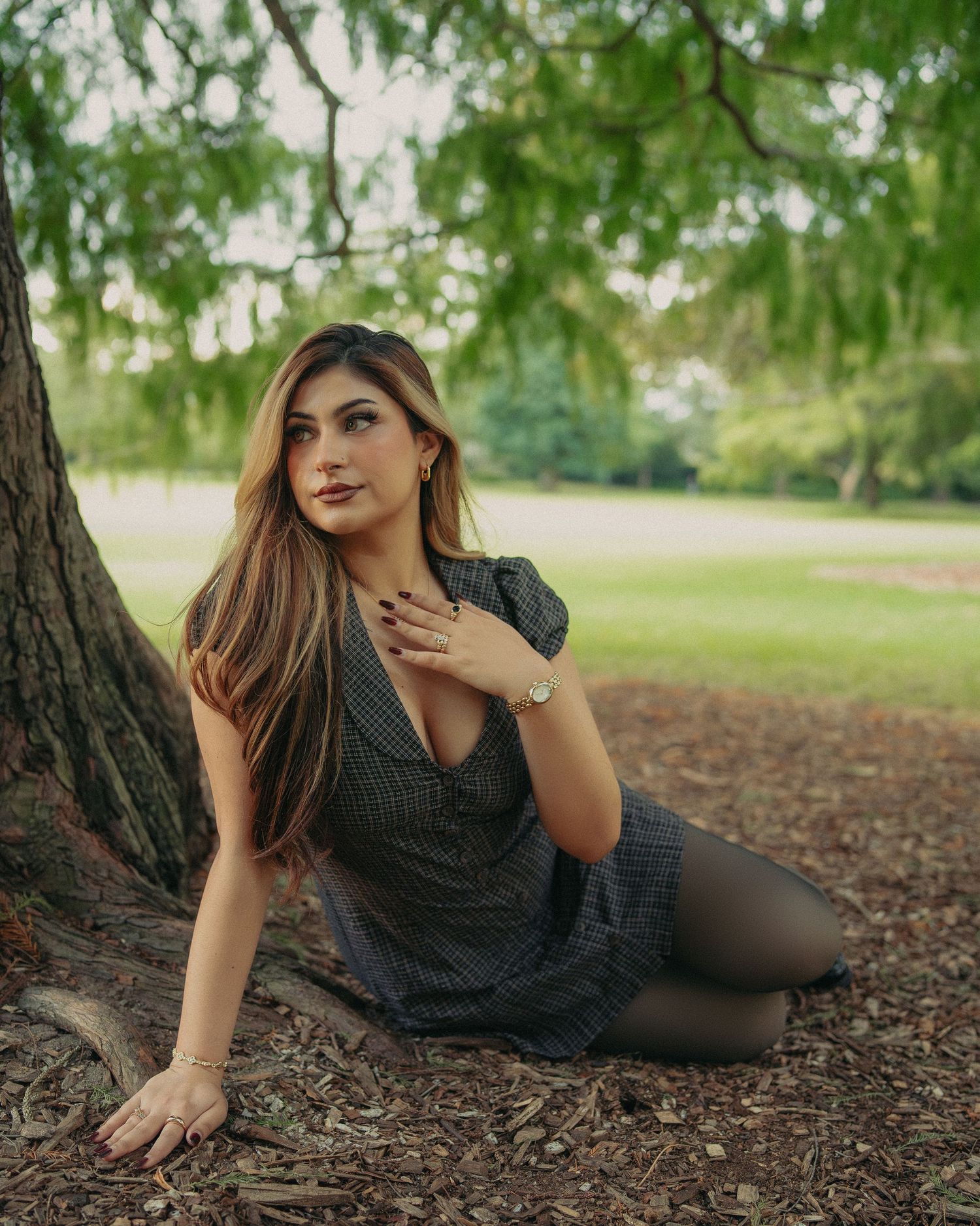 Young woman in plaid dress sitting under a tree in a lush green park.