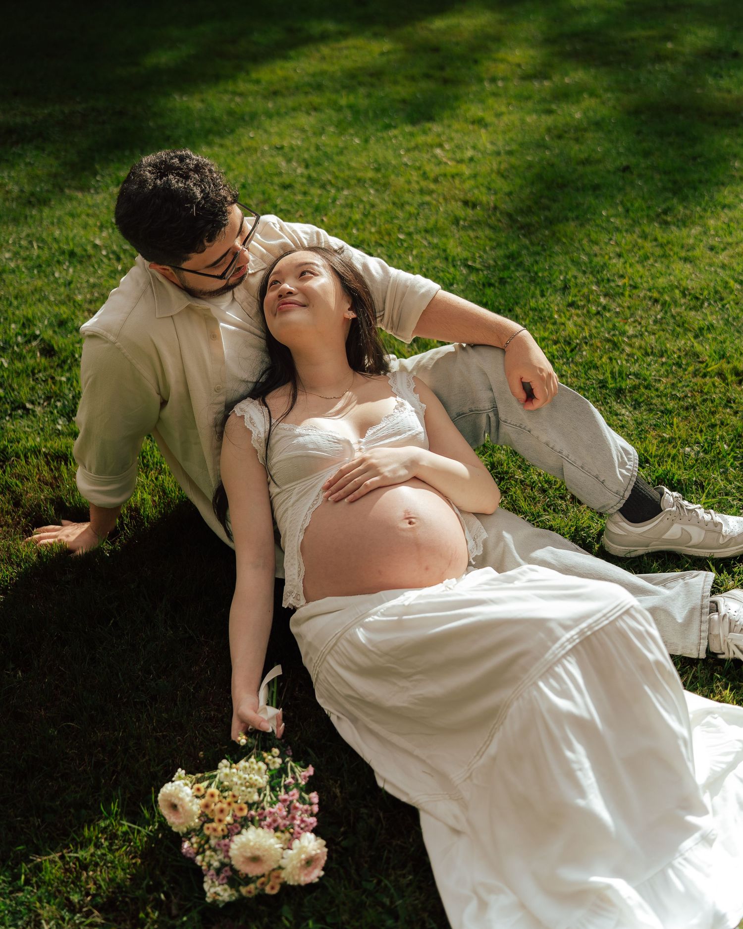 Pregnant woman reclines on grass leaning on partner in dappled sunlight with a bouquet nearby.