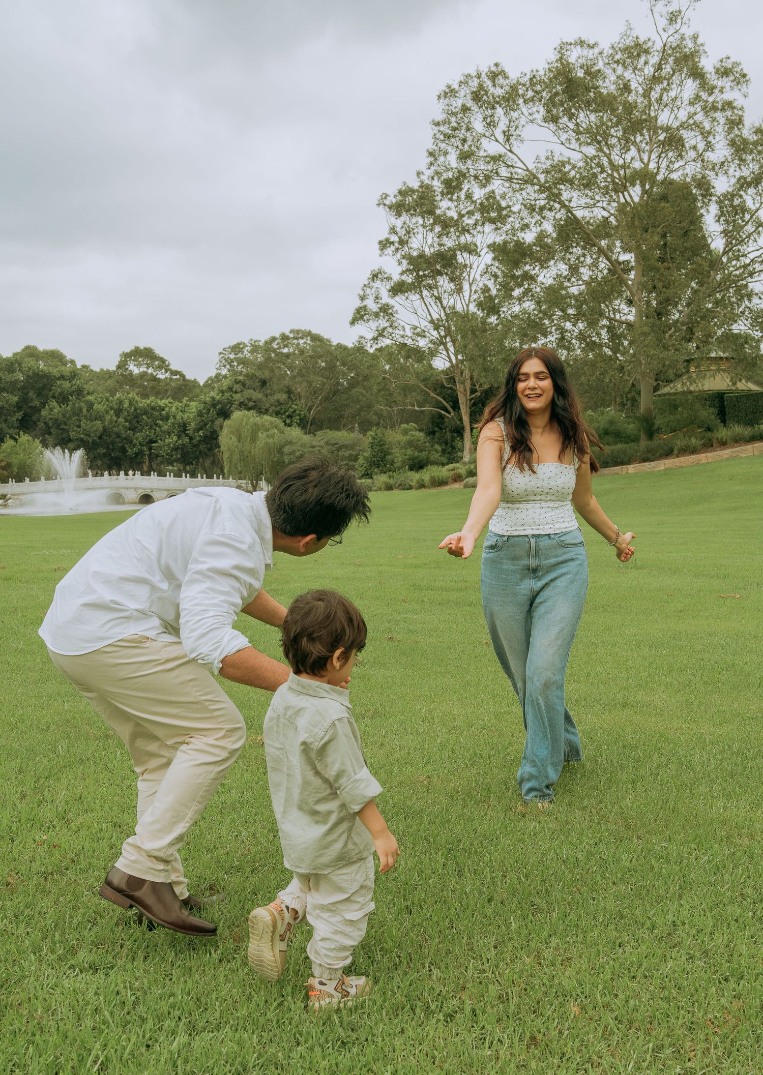 Playful family moment with parents chasing laughing toddler across a green lawn near a fountain.