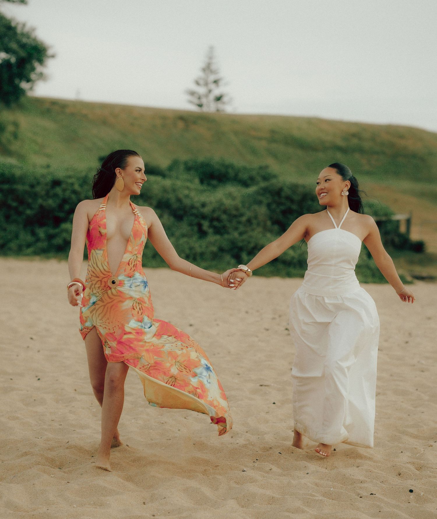 Two women in floral and white dresses holding hands and laughing on a sandy beach.
