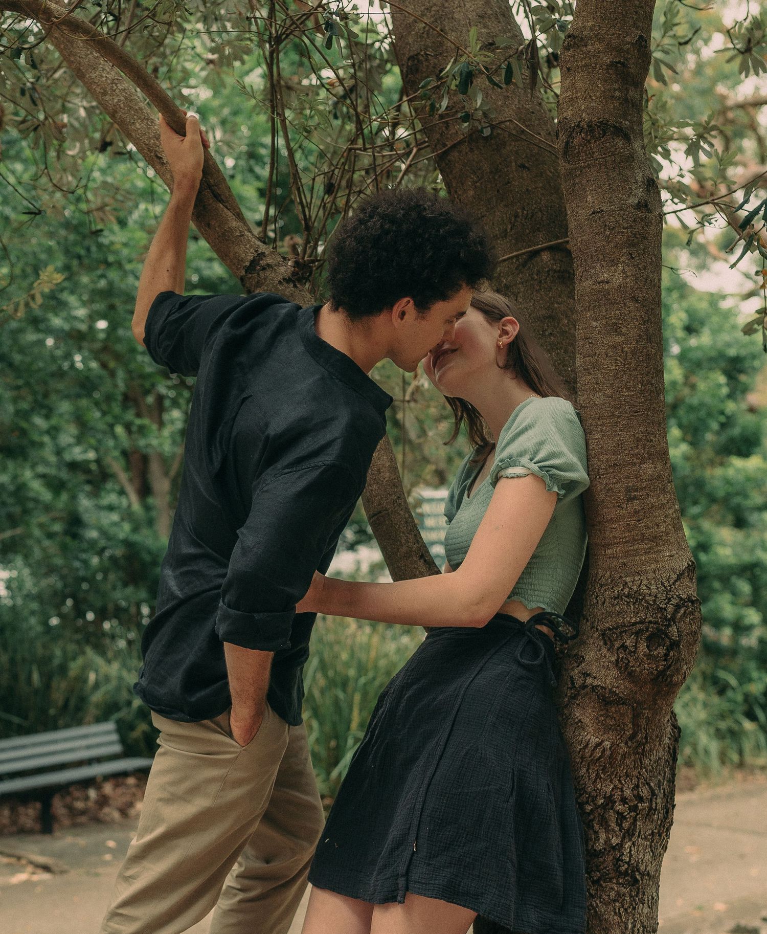 A romantic couple almost kissing while leaning against a tree in a lush green park.