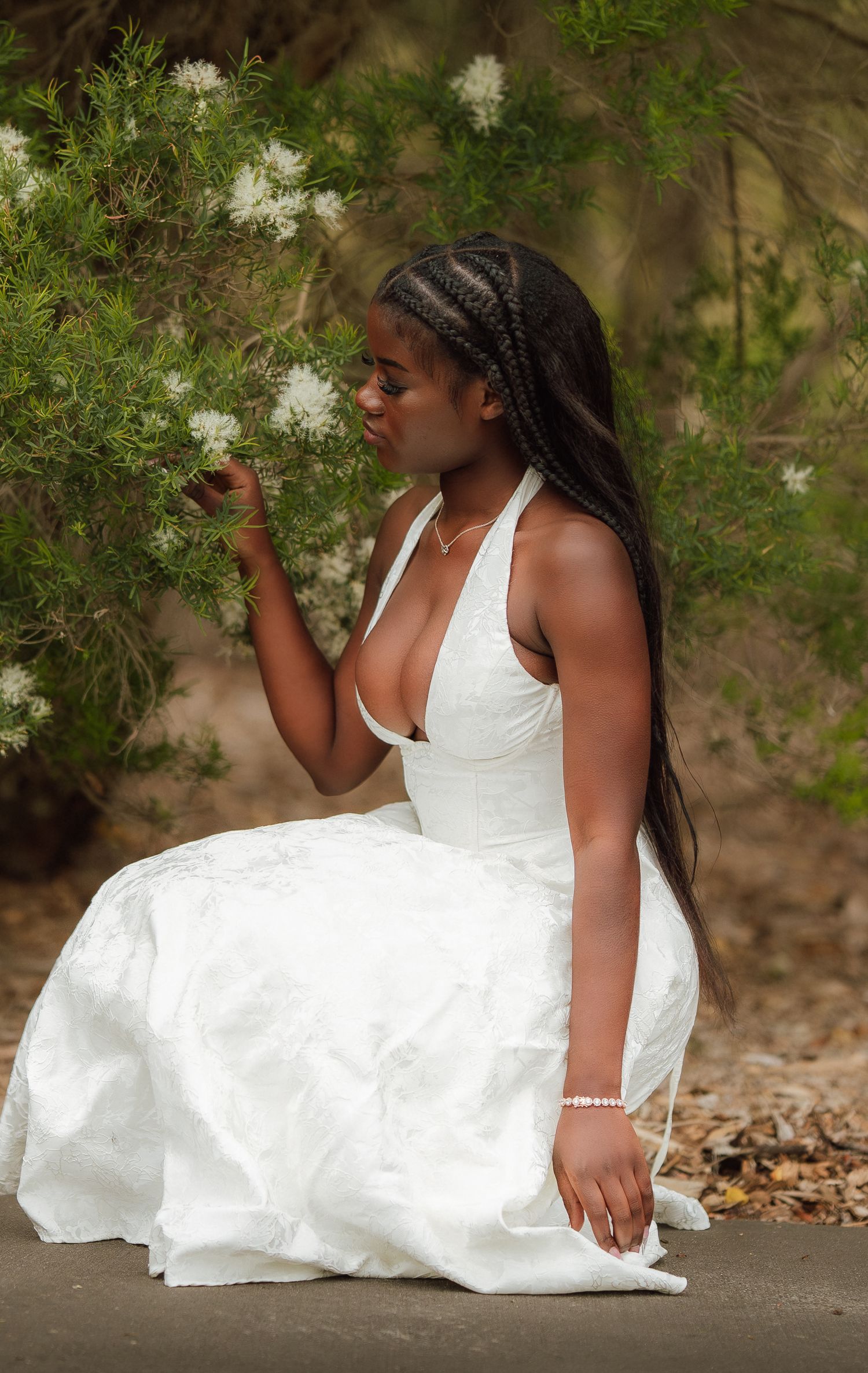 A young Black woman in a white halter dress sits gracefully beside blooming white flowers in a lush garden.