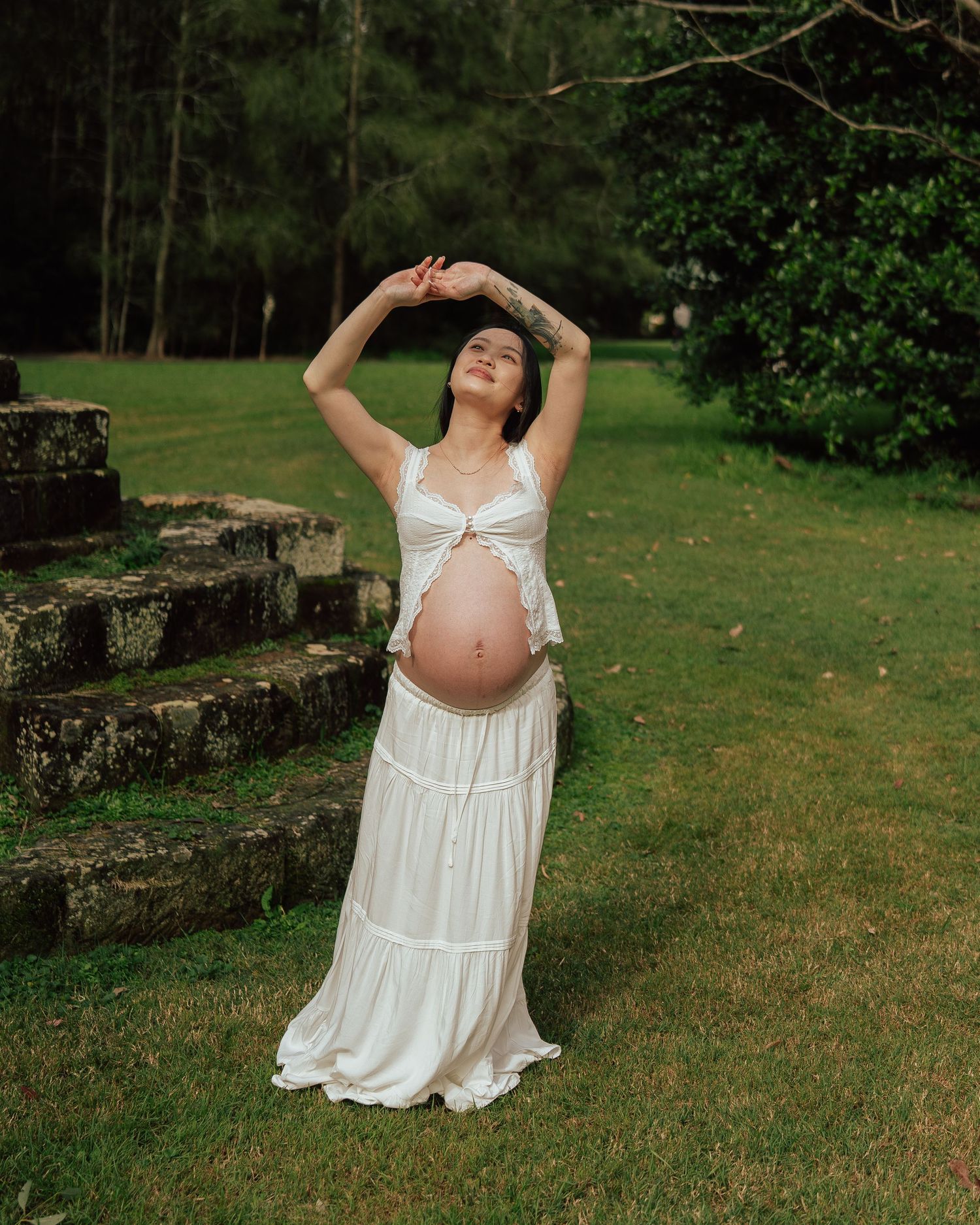 Pregnant woman in white maxi dress posing with arms raised in a serene outdoor garden setting.