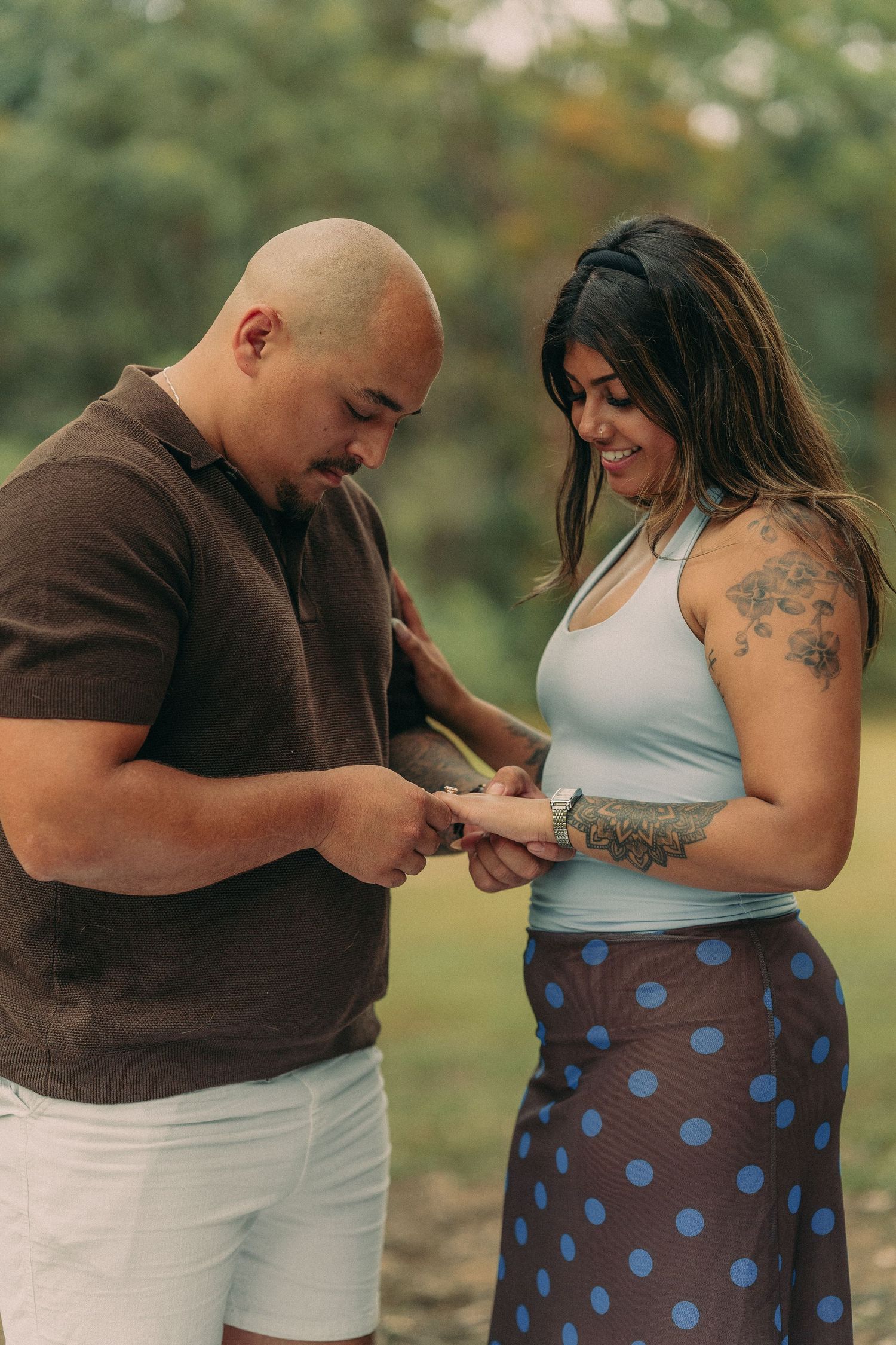 A couple smiles while placing an engagement ring on her finger in a lush green outdoor setting.
