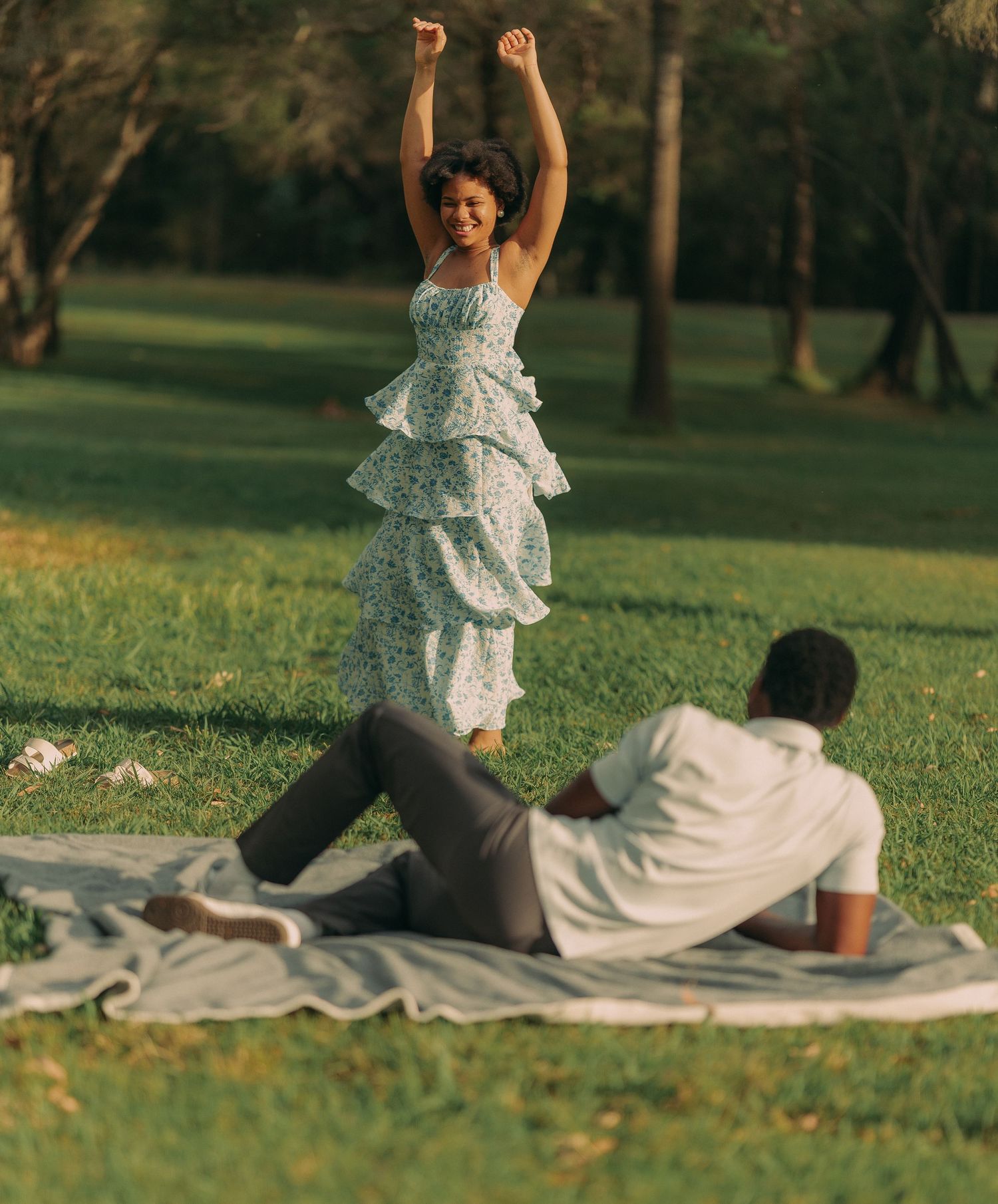 Woman in ruffled floral dress dancing joyfully in a park while man relaxes on a blanket.
