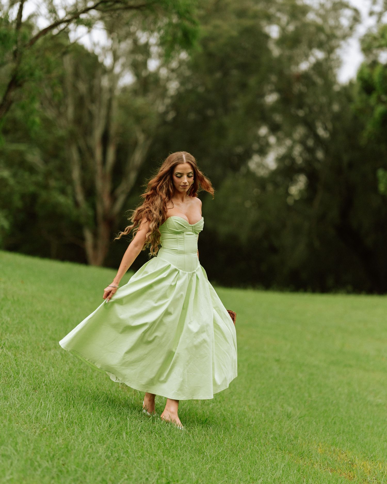 Woman twirling in elegant sage green strapless midi dress on lush green lawn with trees.