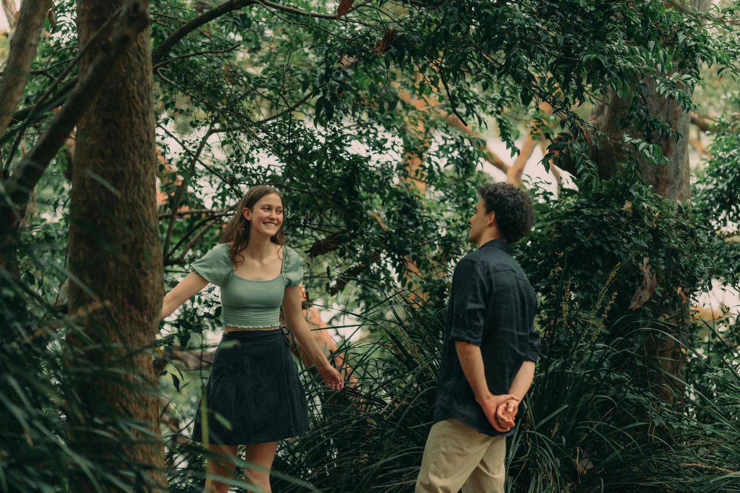 A happy couple walking hand in hand through a lush green forest, smiling at each other.