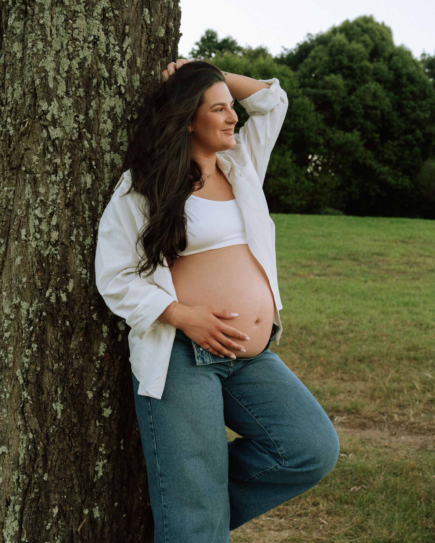 Pregnant woman in white crop top and open shirt leaning against tree, smiling in a lush green park.