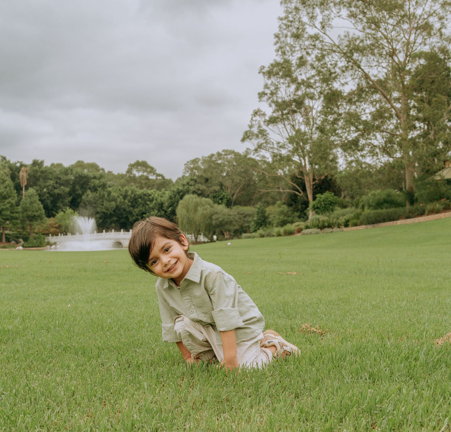Happy young boy in green linen outfit sitting on grass smiling in a scenic park with fountain.