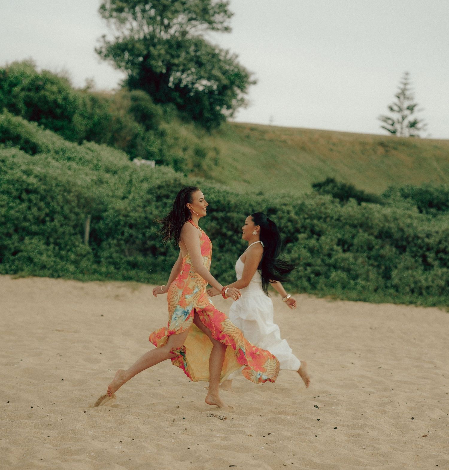 Two women in floral and white dresses dancing joyfully together on a sandy beach.
