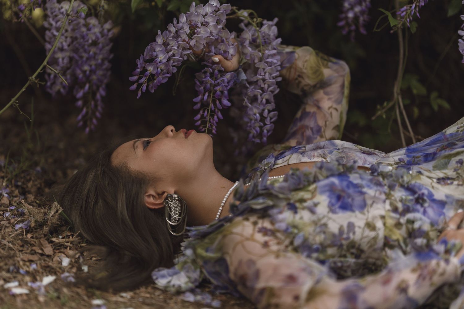 Woman in floral dress lying on ground reaching up to touch purple wisteria flowers.