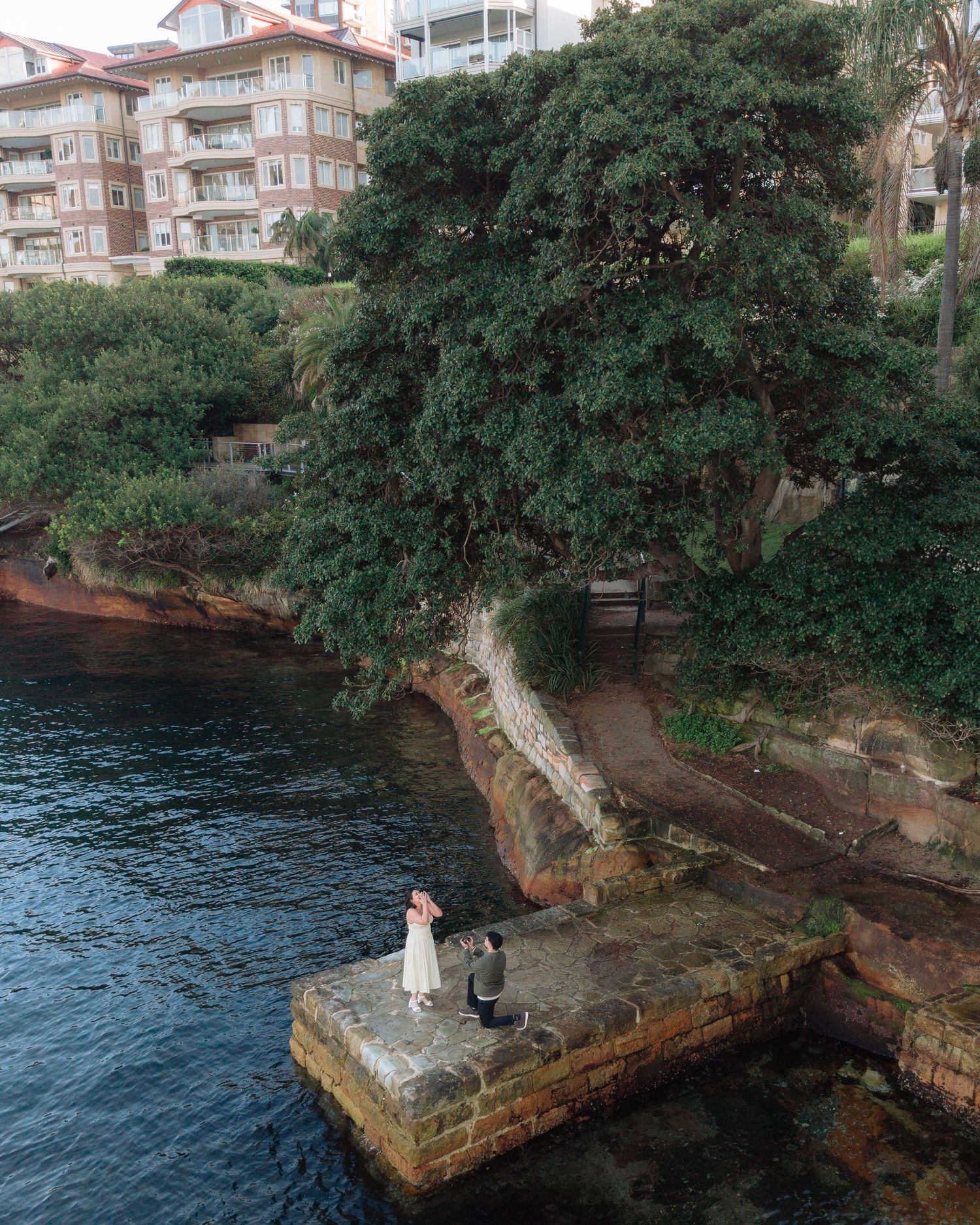 Couple in wedding attire standing on a stone pier surrounded by water, lush trees, and waterfront buildings in Sydney.