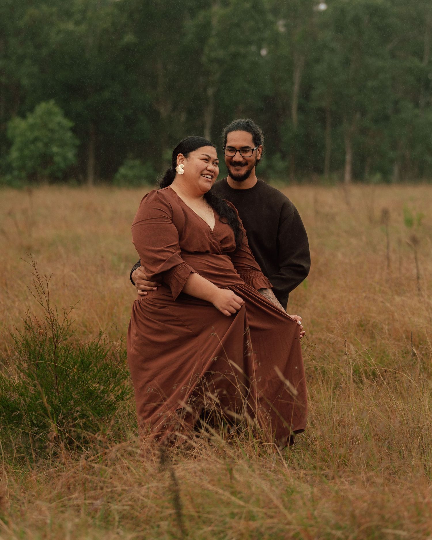 A happy couple laughs together in a golden field, the woman wearing a rust-brown wrap dress.