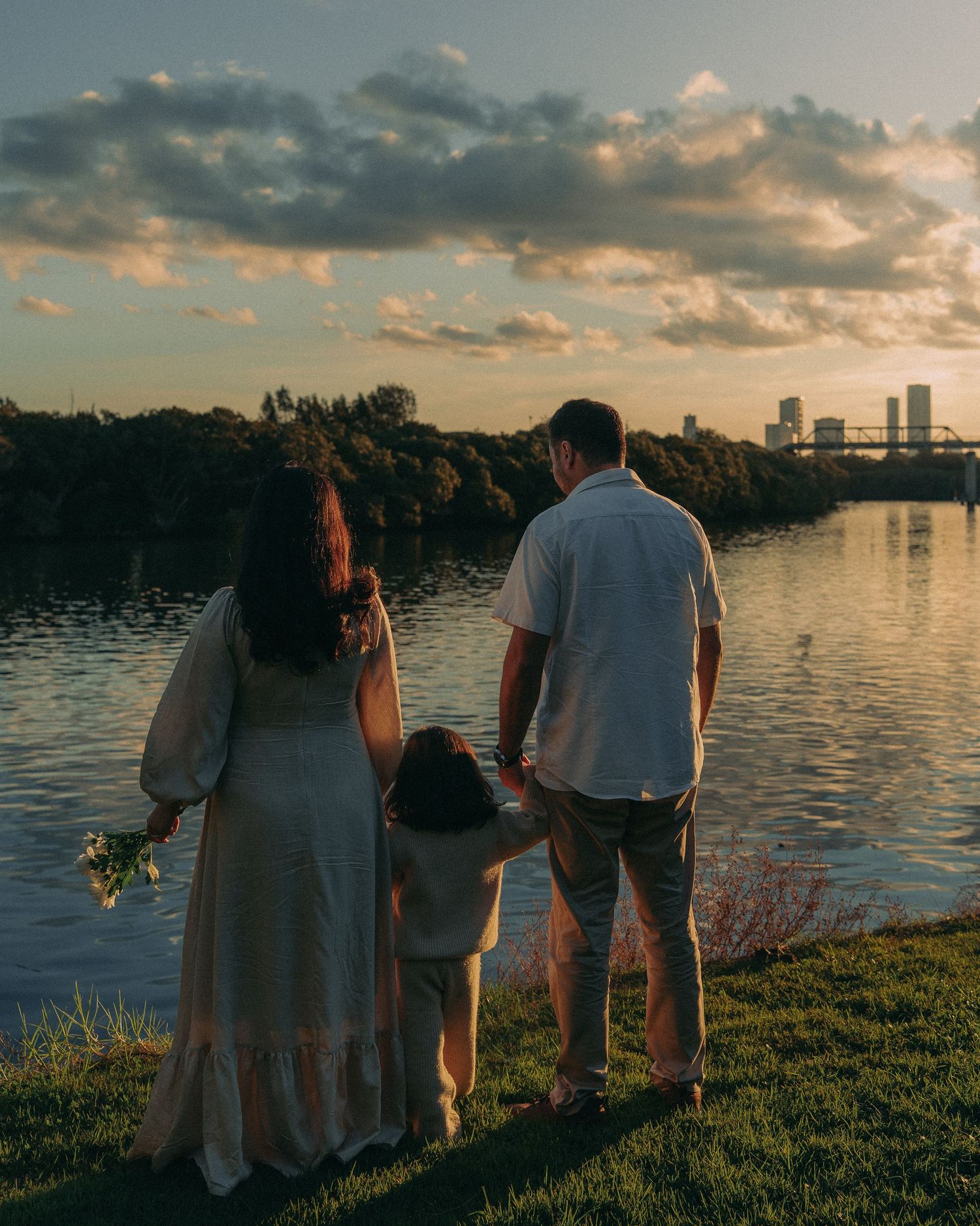 A family of three holds hands while watching a stunning sunset over a lake with a city skyline in the distance.