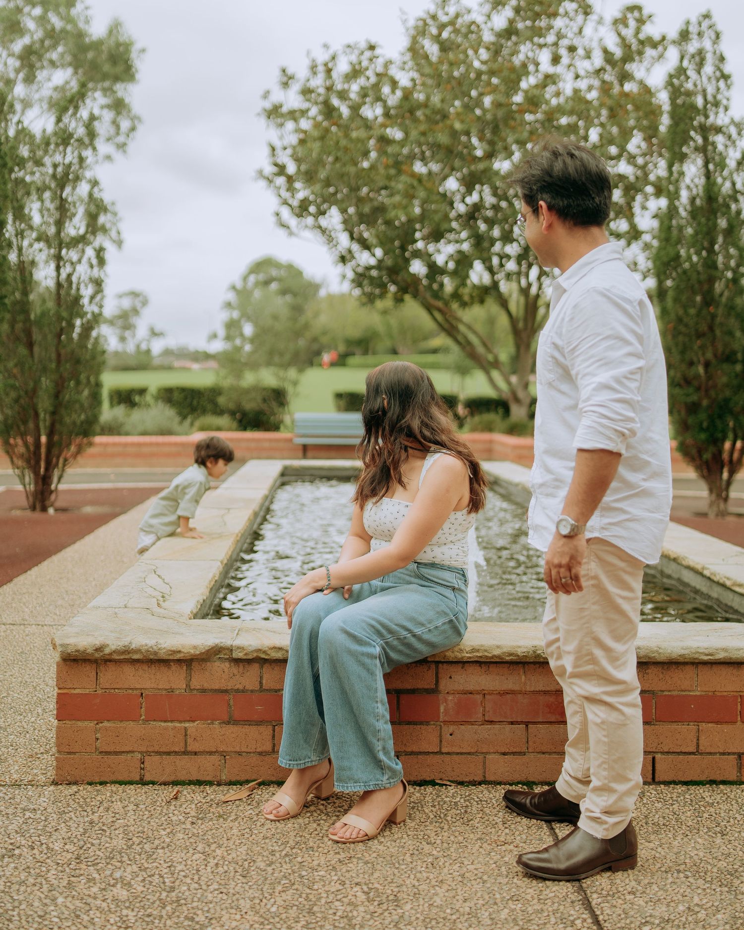 Woman sitting on brick ledge beside garden fountain while man stands nearby watching young child play.