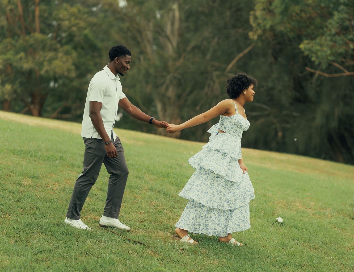 A couple walks hand in hand across a green grassy park, the woman in a tiered floral dress.
