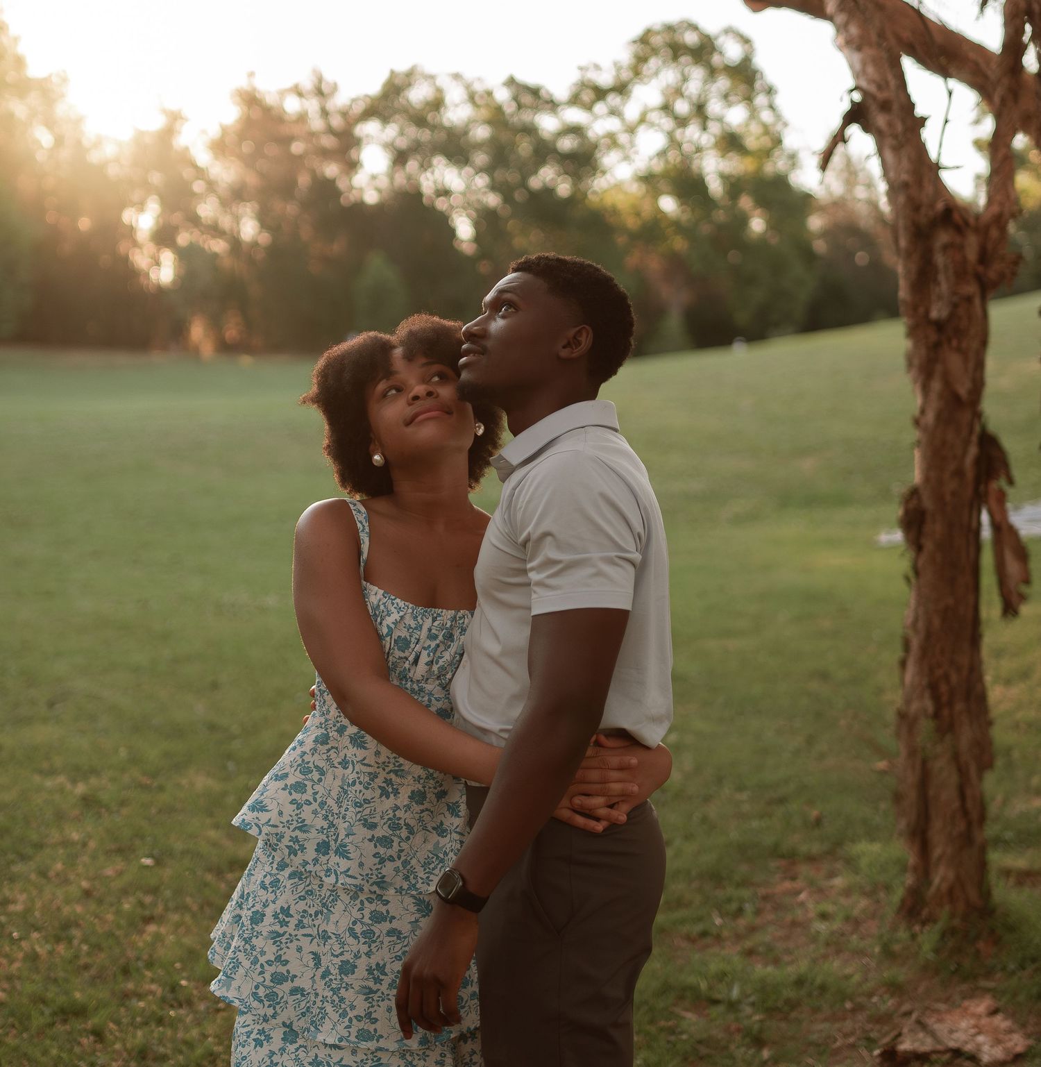 A loving couple embraces and gazes upward together near a tree in a park at golden sunset.