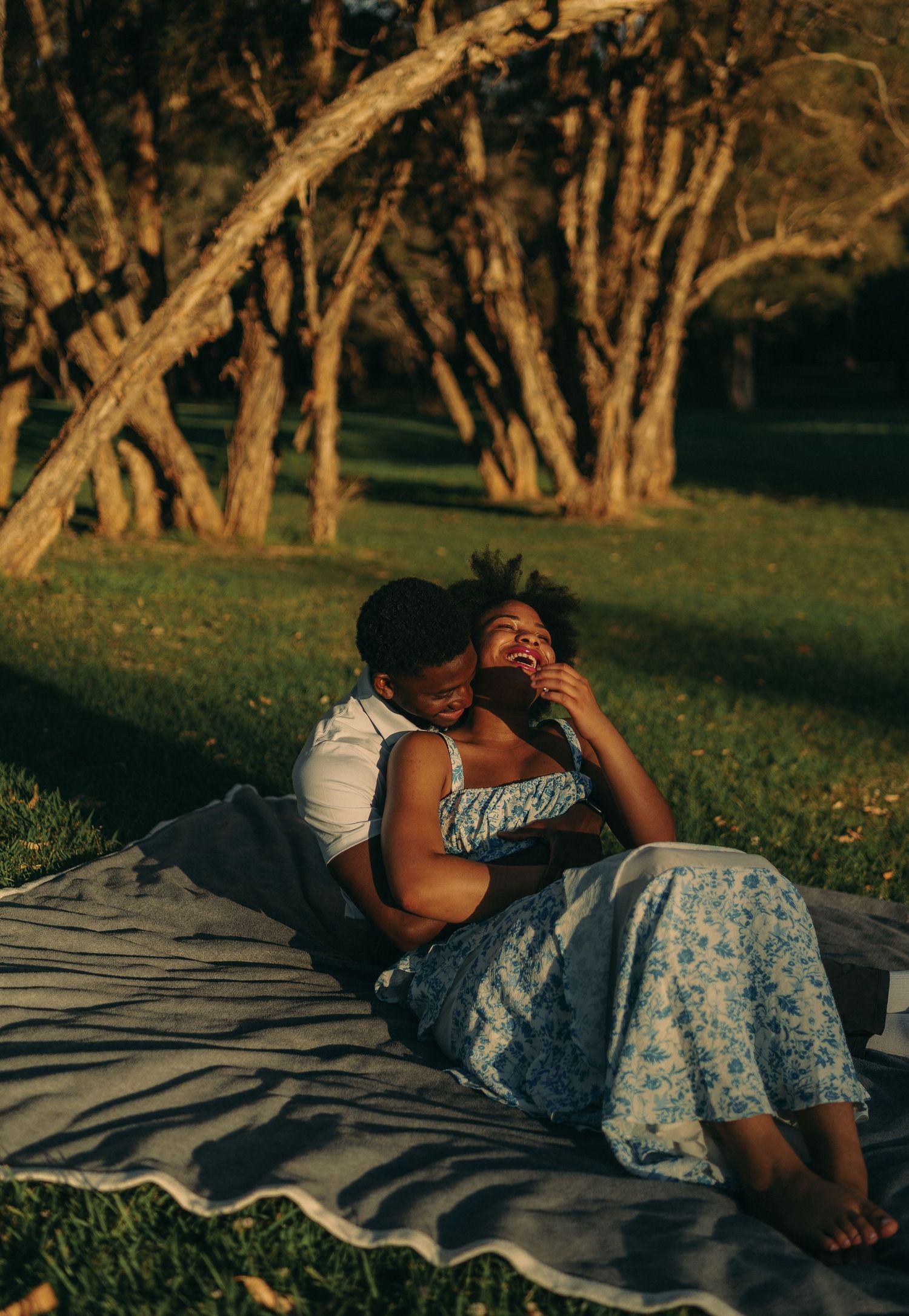A happy couple cuddles and laughs together on a blanket in a sunlit park among trees.