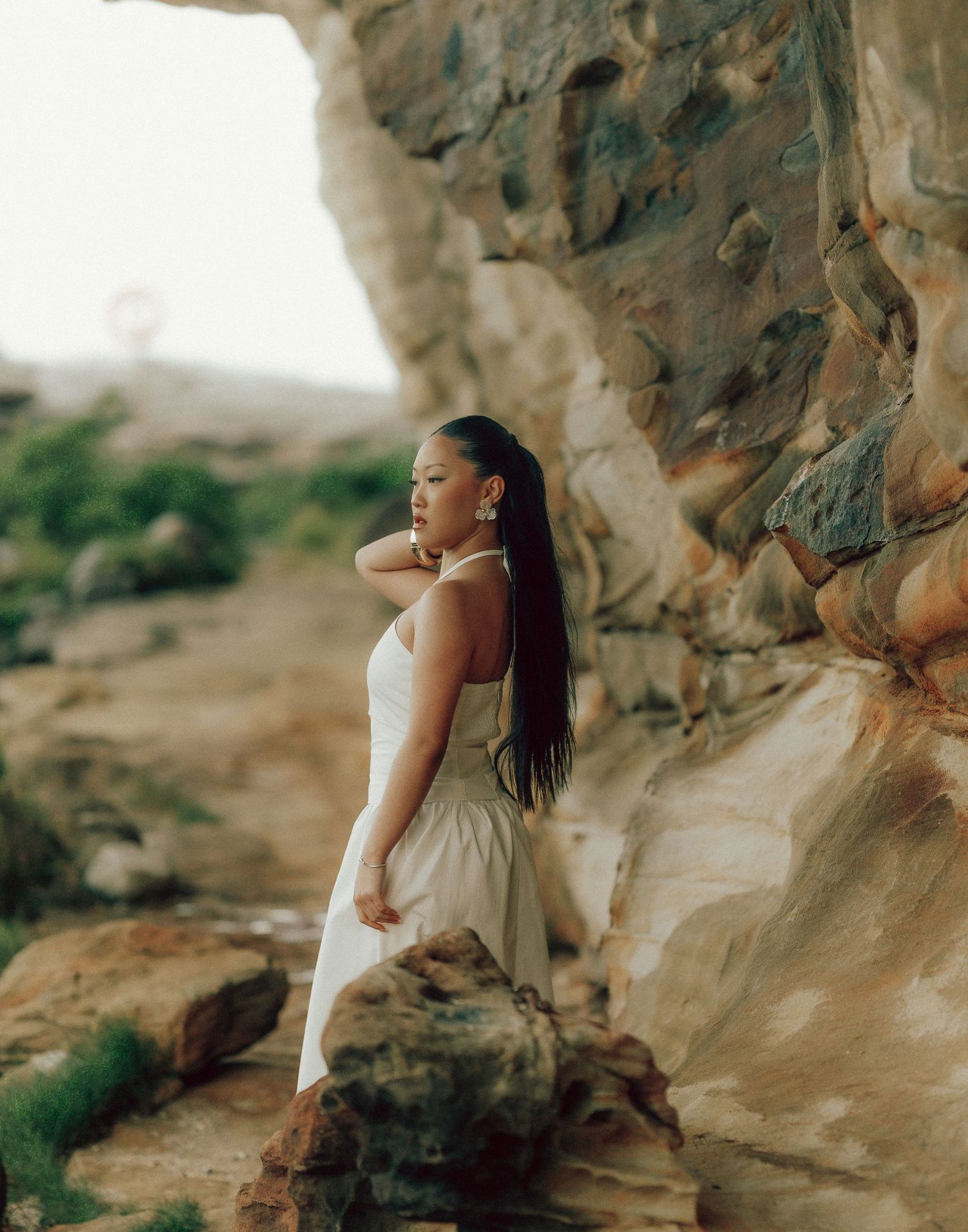 Asian woman in white halter dress posing beside large sandstone rock formation in natural outdoor setting.