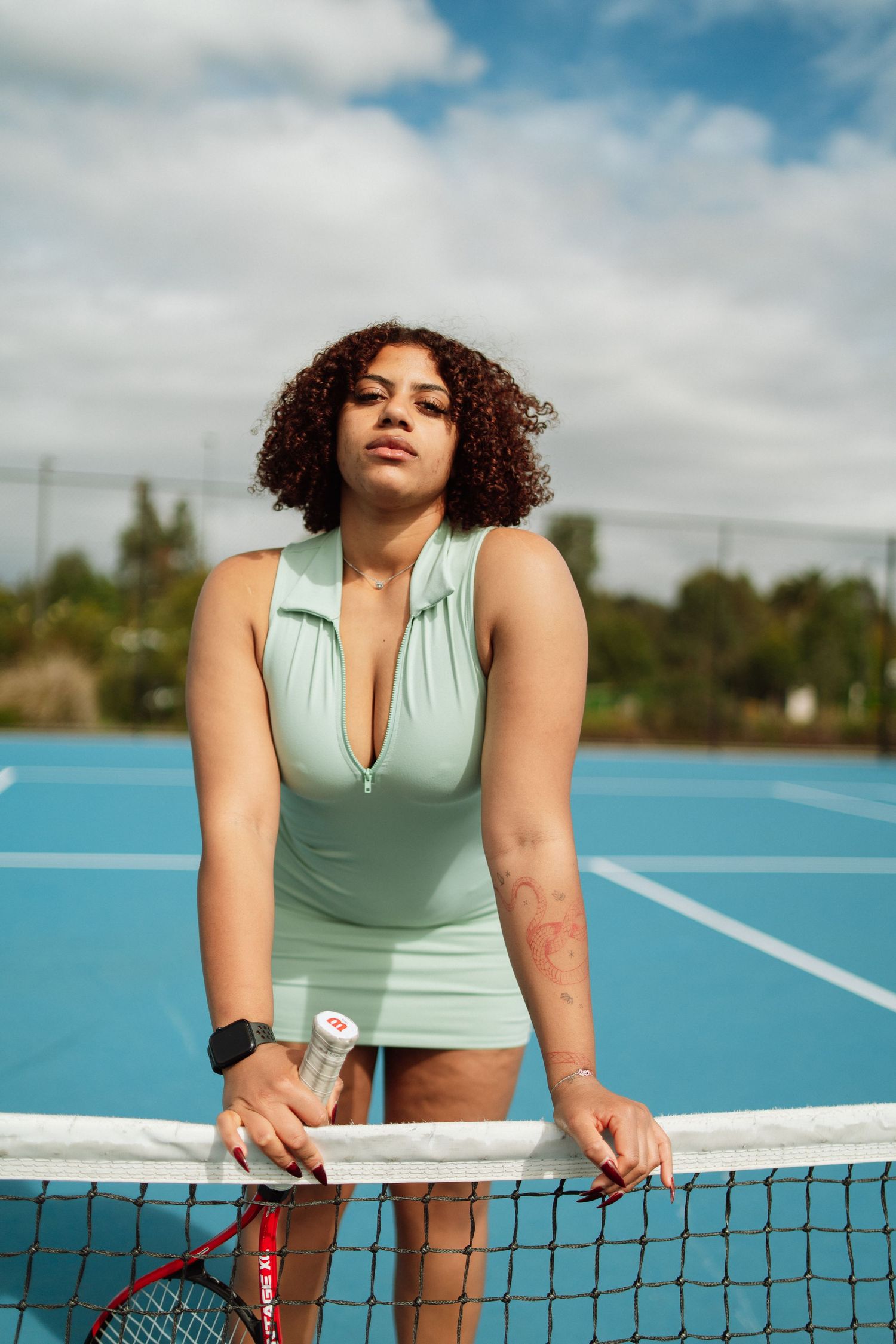 A woman in a mint green tennis dress leans on the net holding a racket on a sunny outdoor blue court.