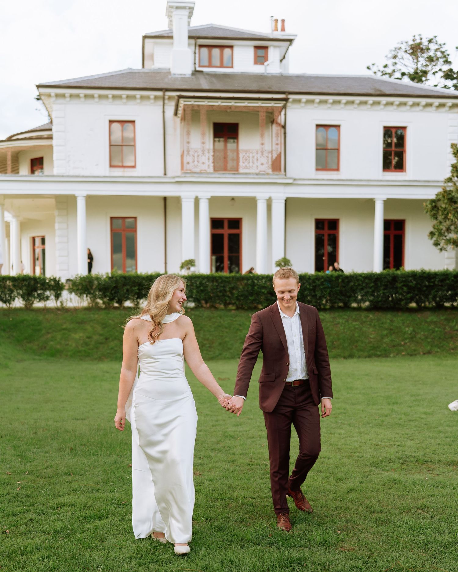 Smiling bride and groom in white gown and burgundy suit holding hands on lawn of grand estate.