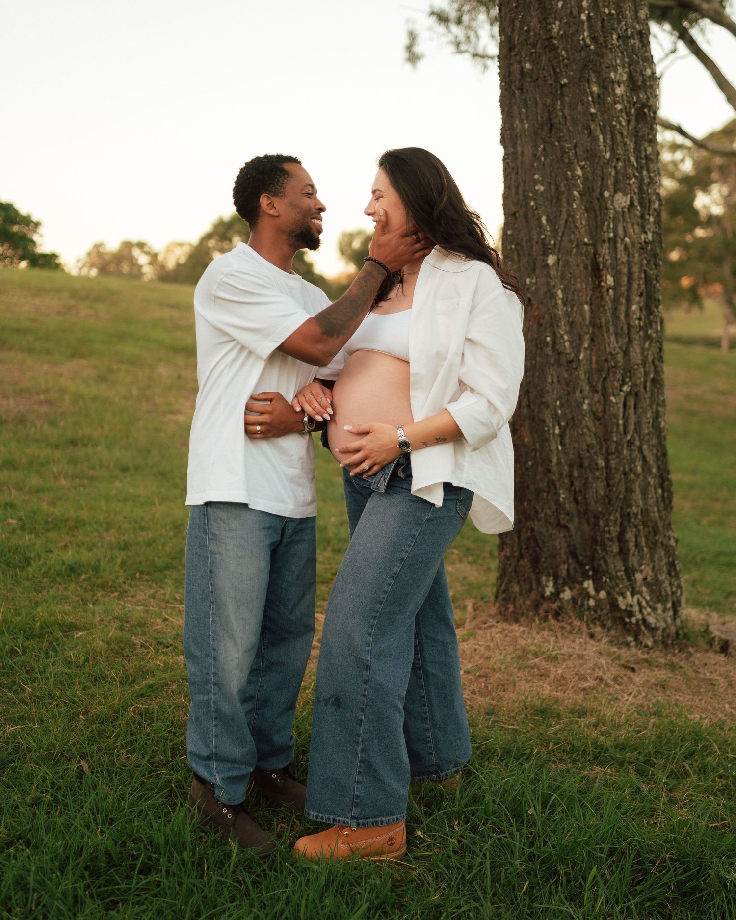 Couple in matching white shirts and jeans share a joyful moment outdoors, showcasing a beautiful baby bump during maternity photos.