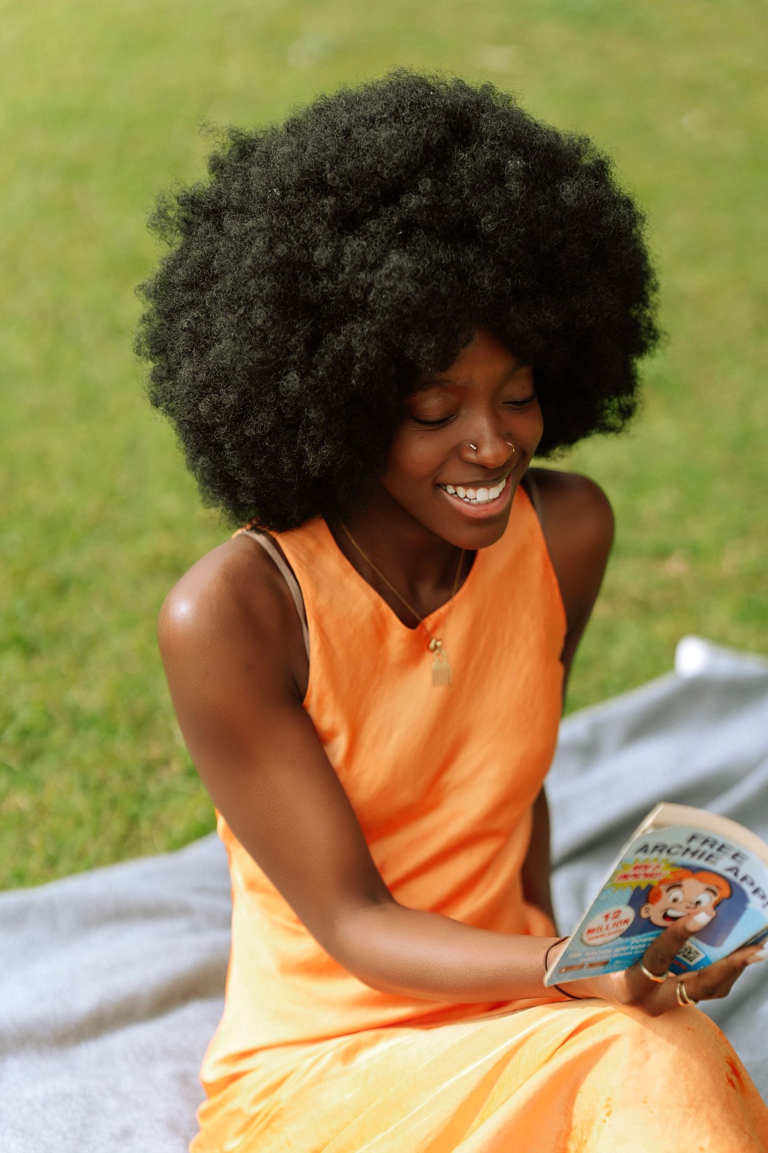 Joyful woman with afro hairstyle in orange dress reads a book while sitting on blanket in park.