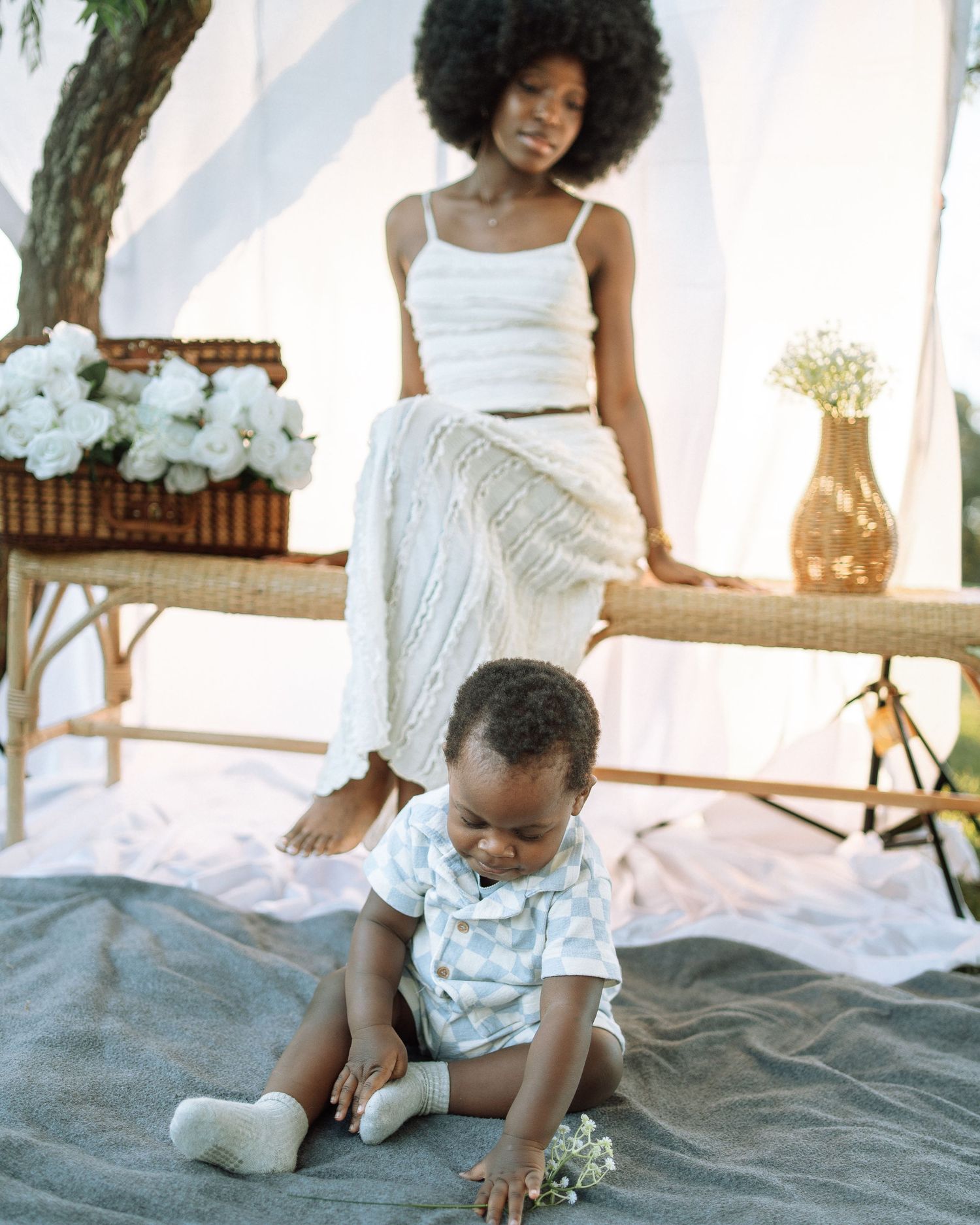 A woman in white sits on a rattan bench while her baby plays on a blanket in an outdoor picnic setting.