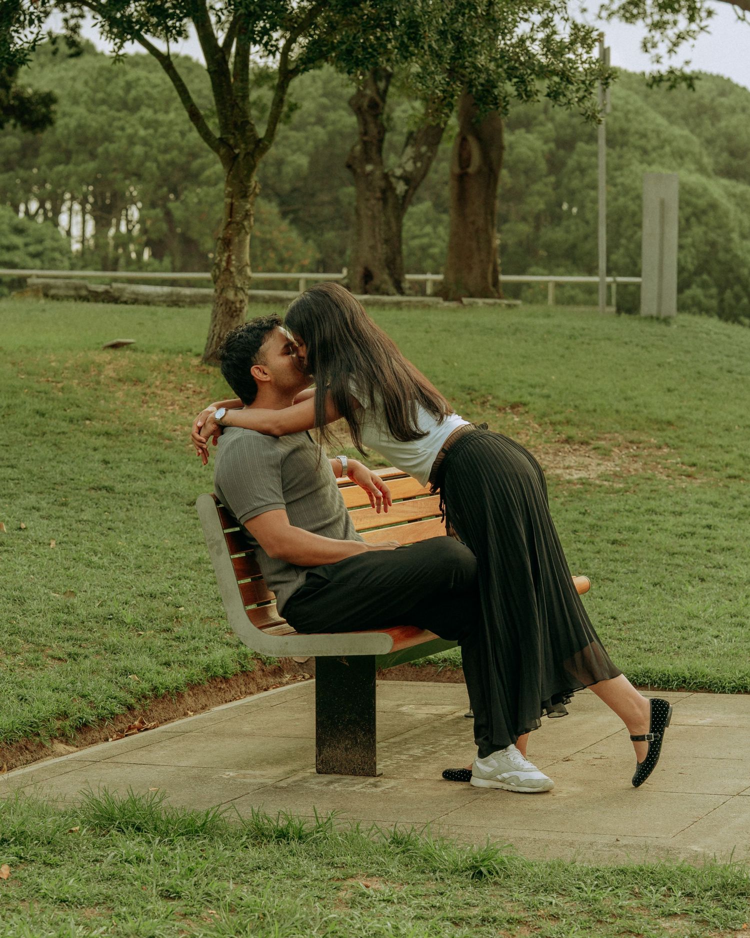 Couple sharing a tender kiss while sitting together on a wooden park bench surrounded by trees.