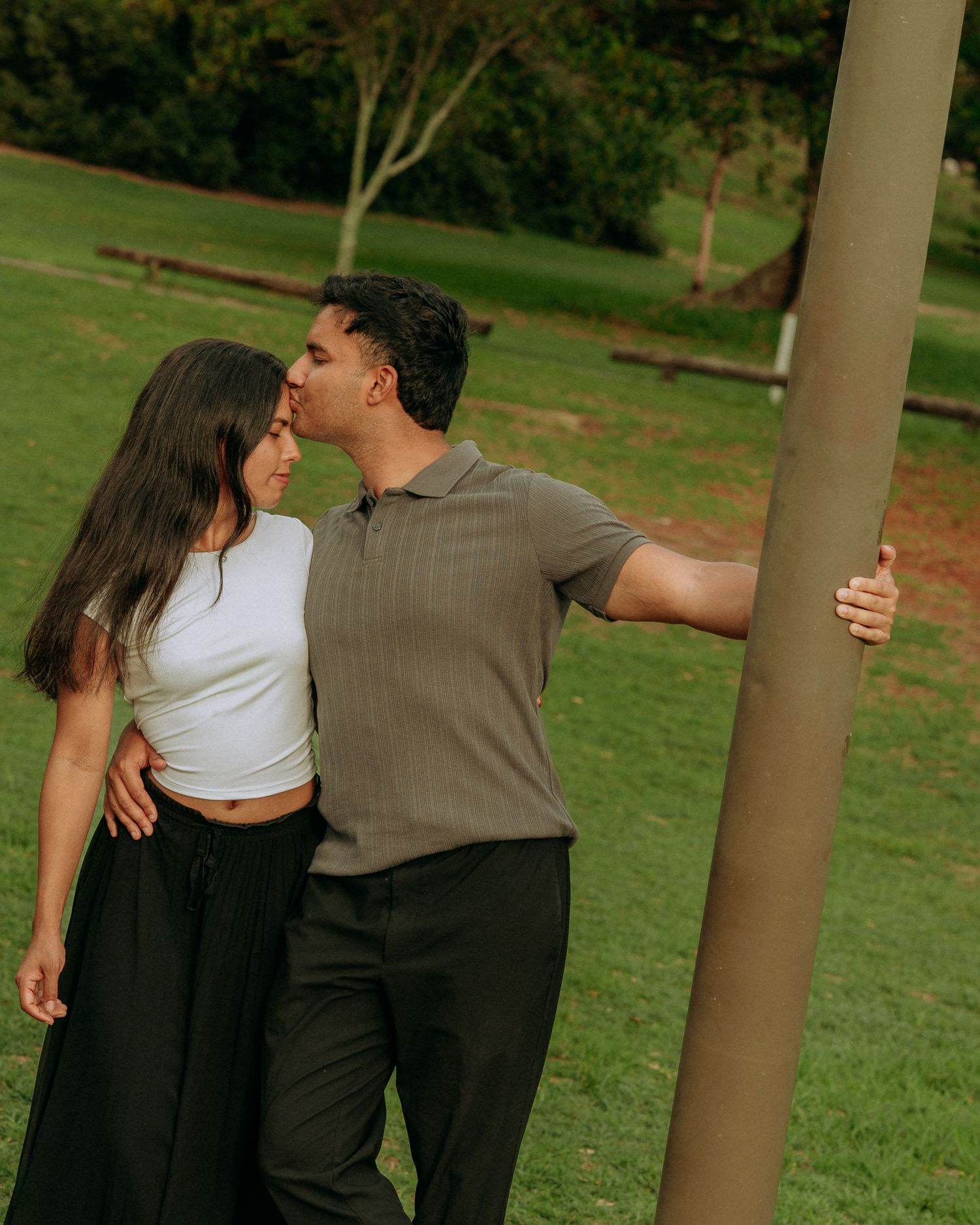 A couple shares a tender kiss in a lush green park, the man in a grey polo embracing a woman in a white crop top.