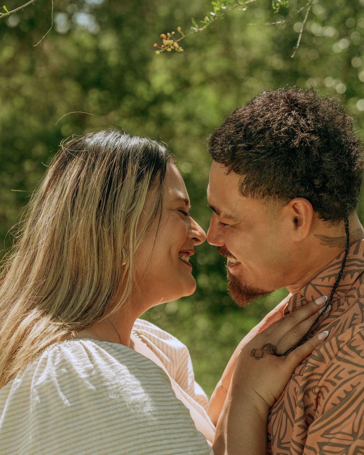Couple smiling and leaning in close together outdoors surrounded by lush green foliage in sunlight.