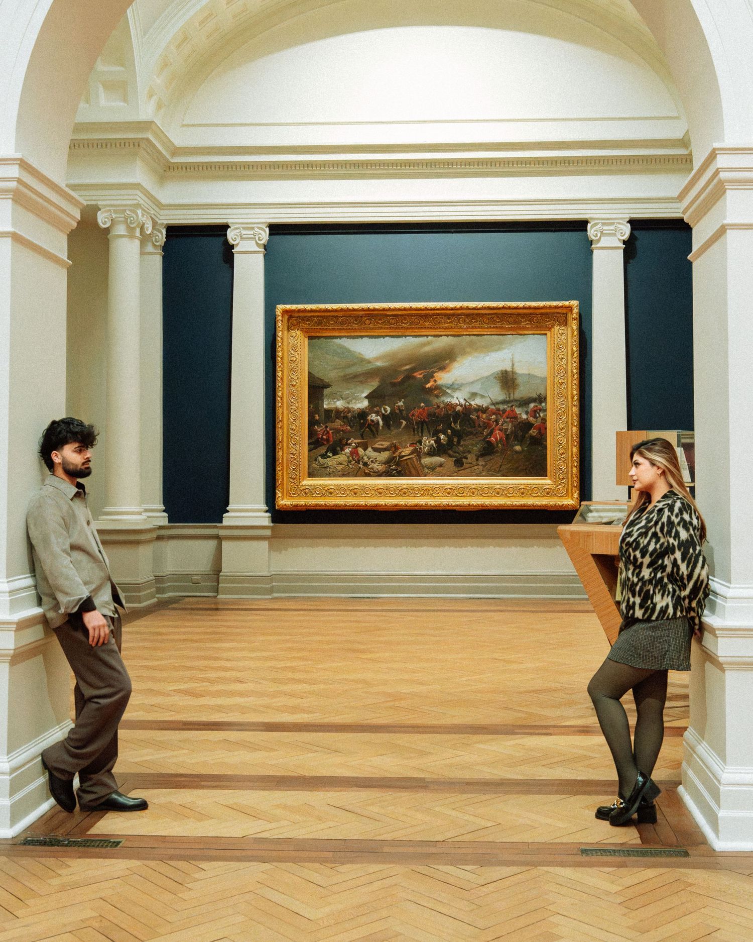 Man and woman standing apart in a grand art gallery beside a large gold-framed painting.