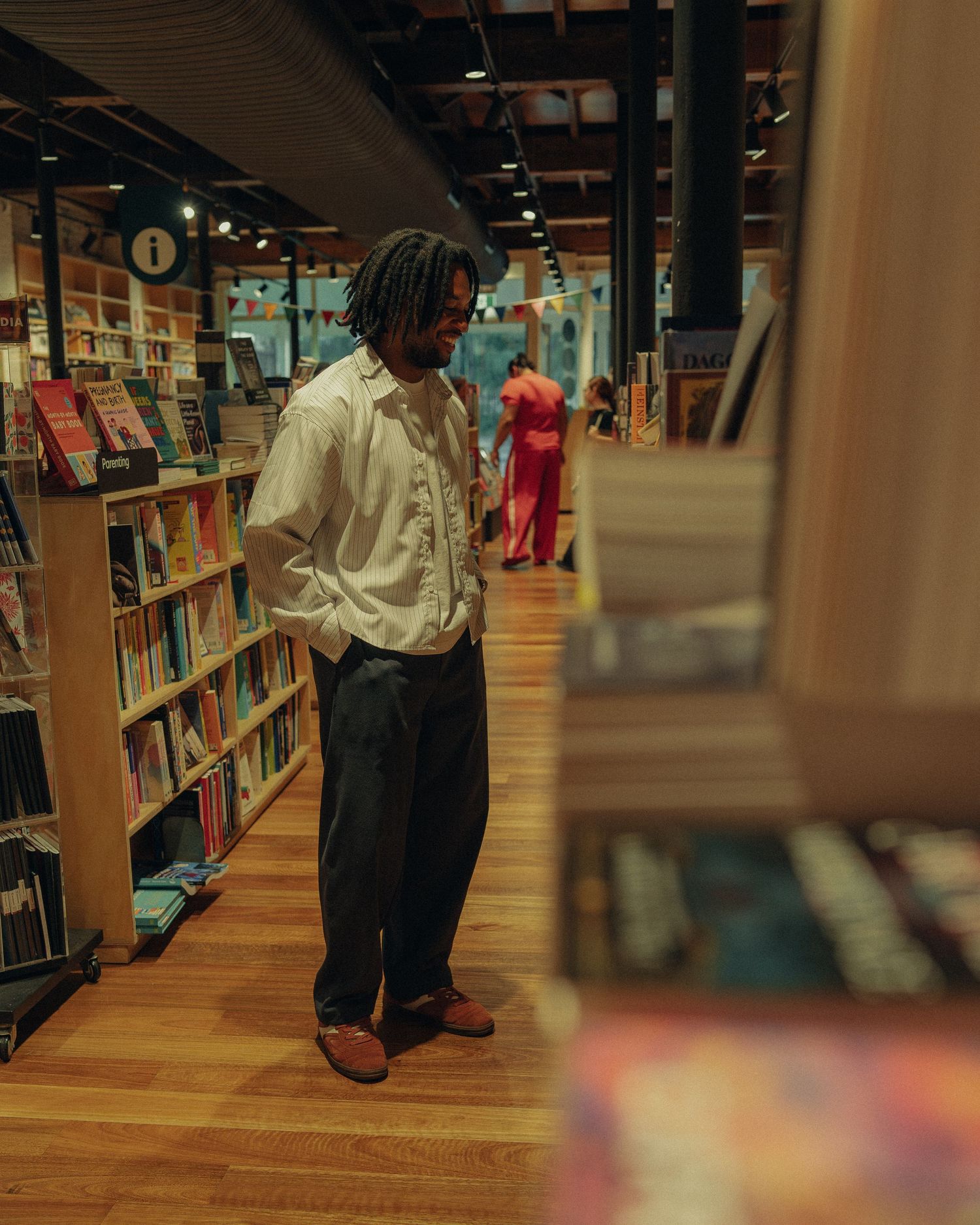A man with dreadlocks browses books in a warmly lit bookstore with wooden floors.