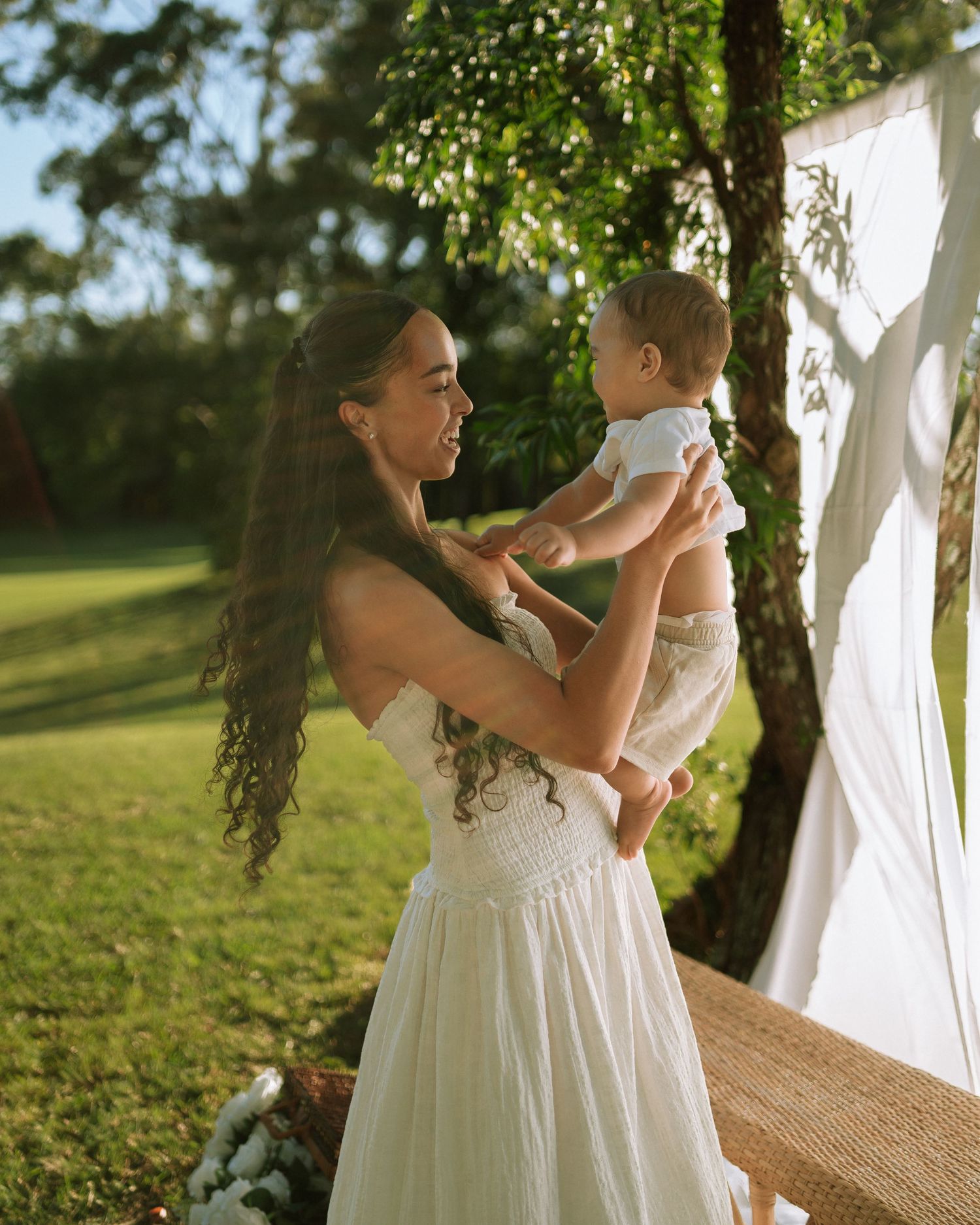 A woman in a white dress holds her baby outdoors in a sunlit garden with green grass.