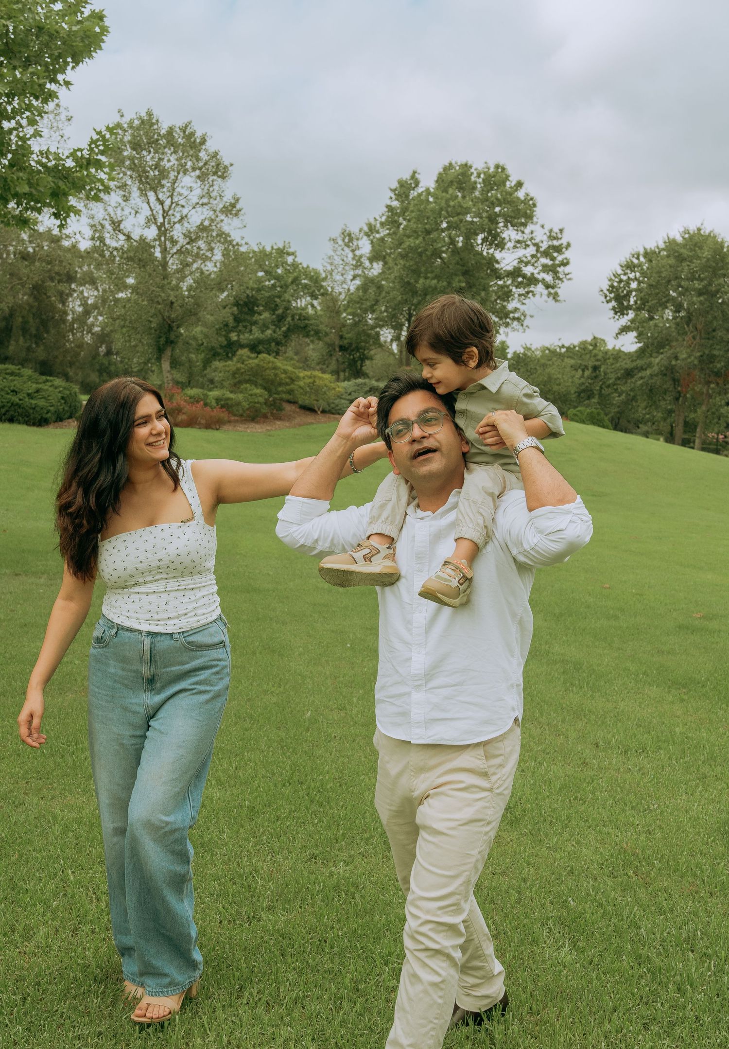 A joyful family walks through a green park, father carrying child on shoulders while mother smiles nearby.
