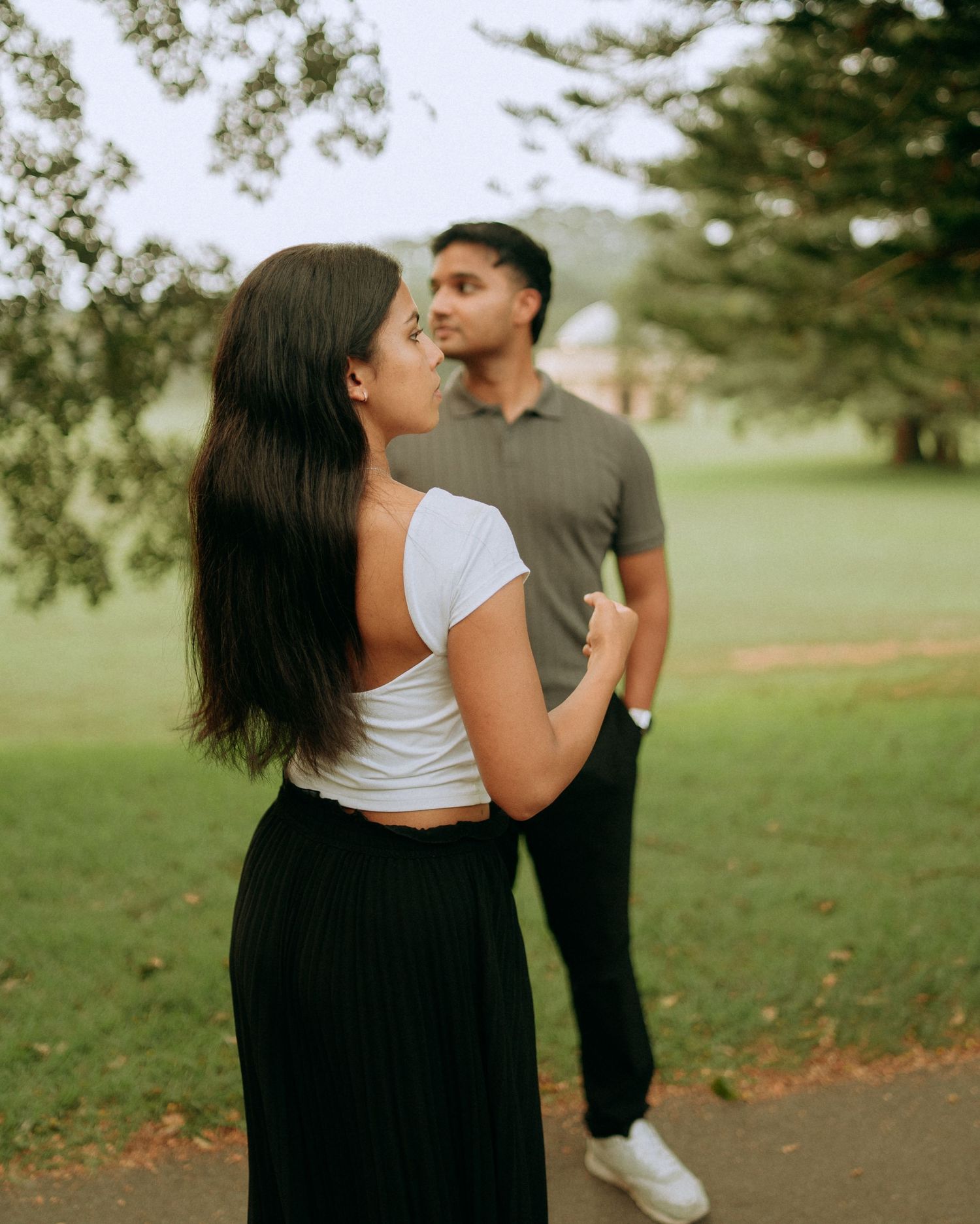 A woman in a white crop top and black skirt looks over her shoulder at her partner standing behind her in a park.