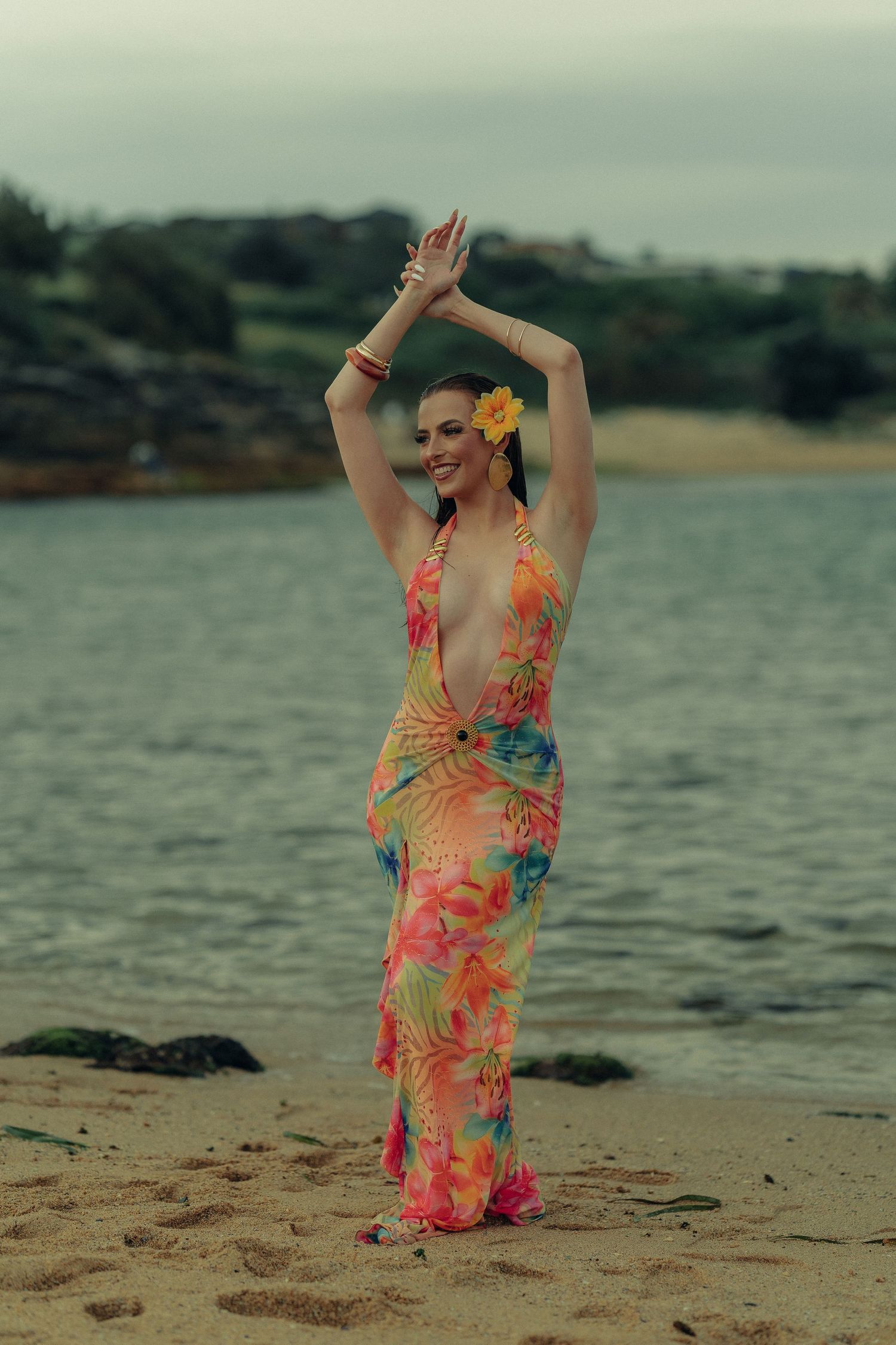 Woman in colorful floral halter dress posing with arms raised overhead on sandy beach at twilight.