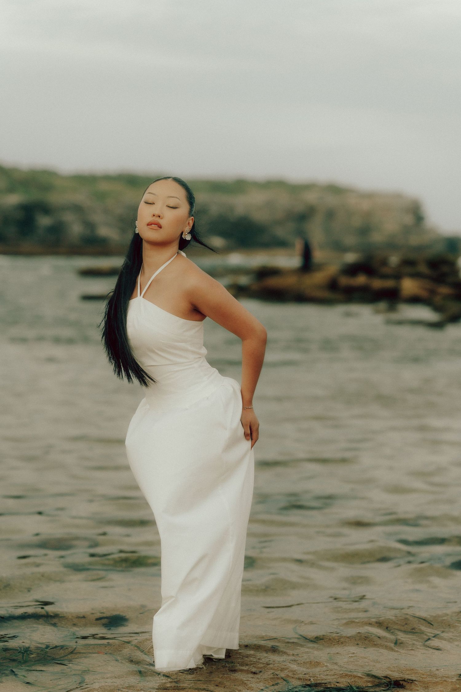 Woman in white halter maxi dress posing gracefully on wet sandy shore with rocky coastline behind.