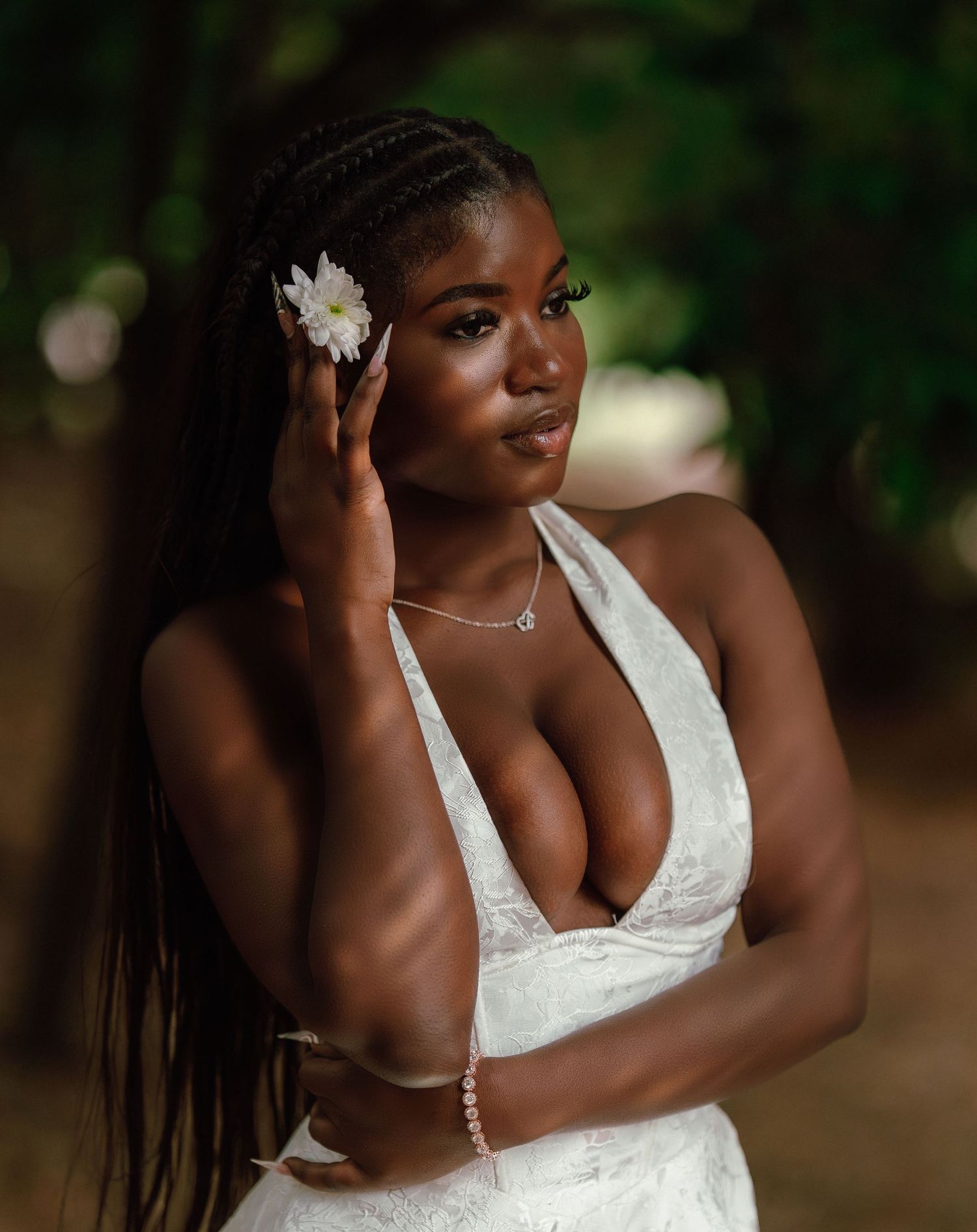 Young Black woman in white halter dress with braided hair and white flower poses in green setting.