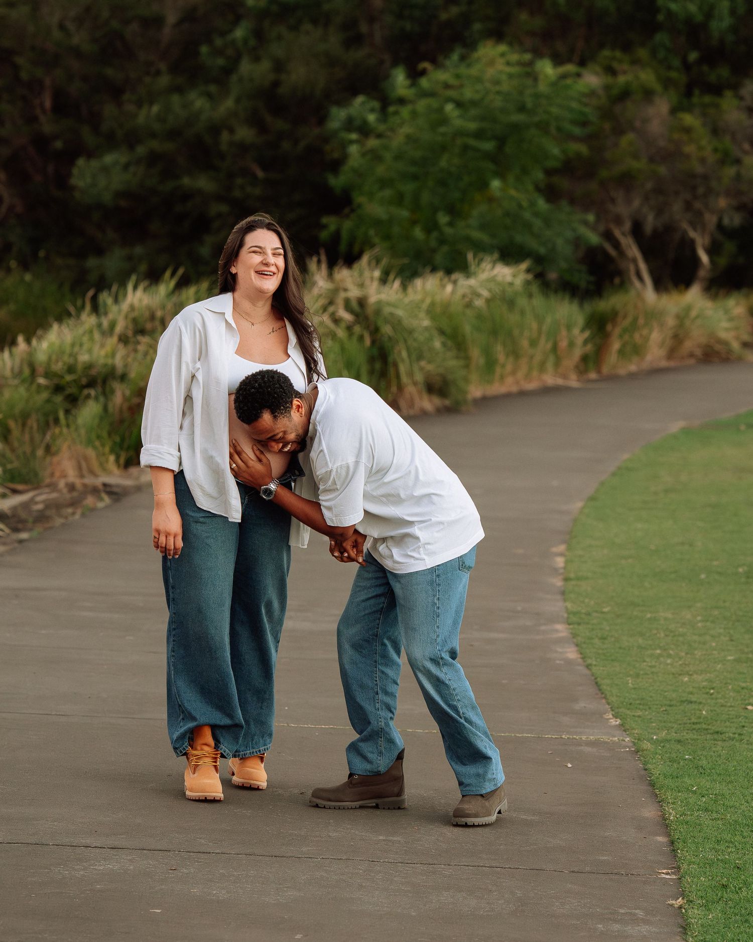Expecting couple in white shirts laughing together on a curved pathway surrounded by lush greenery.