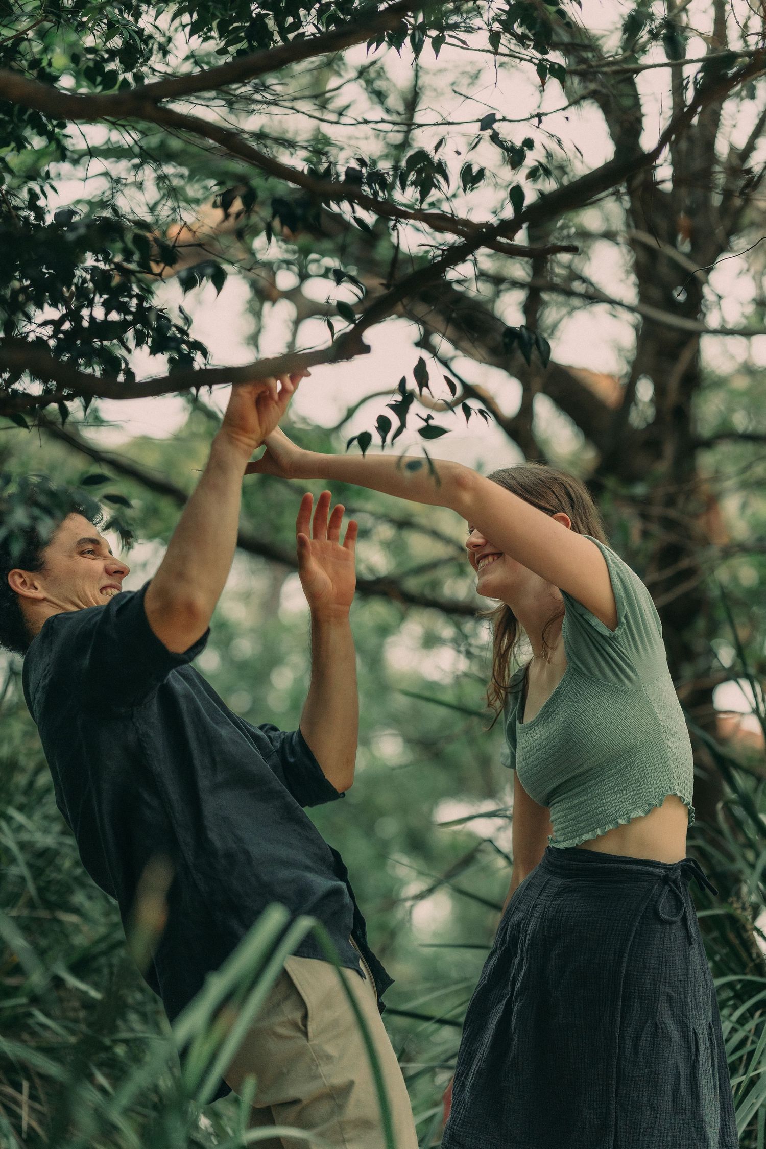 A playful couple reaching up together toward tree branches in a green outdoor setting.